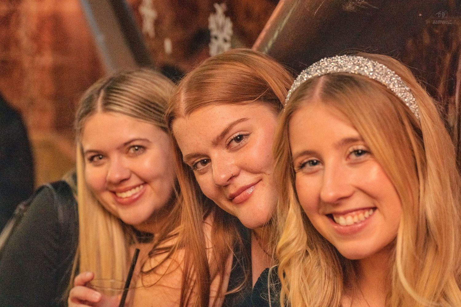 Three smiling young women posing together at a nightclub, one wearing a sparkly headband.
