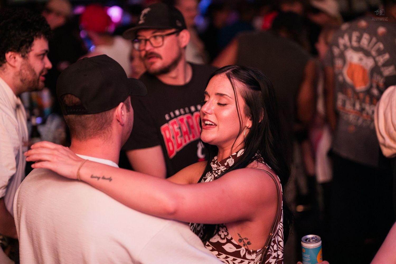 Young woman with tattoos embraces a man in a black cap on a busy nightclub dance floor.