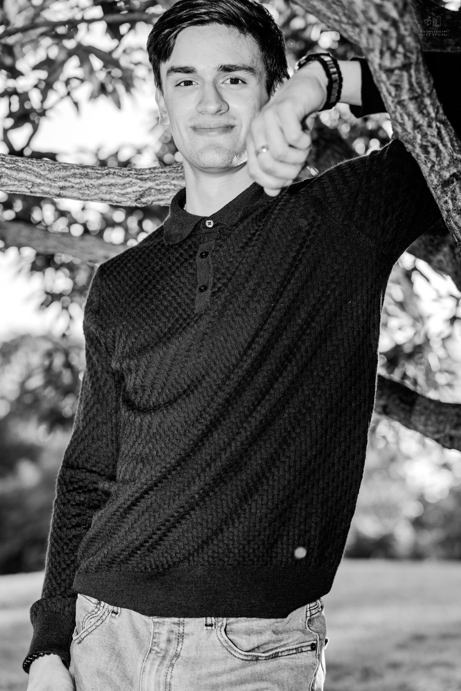Young man in dark polo sweater and jeans leaning against a tree branch, smiling outdoors in black and white photo.