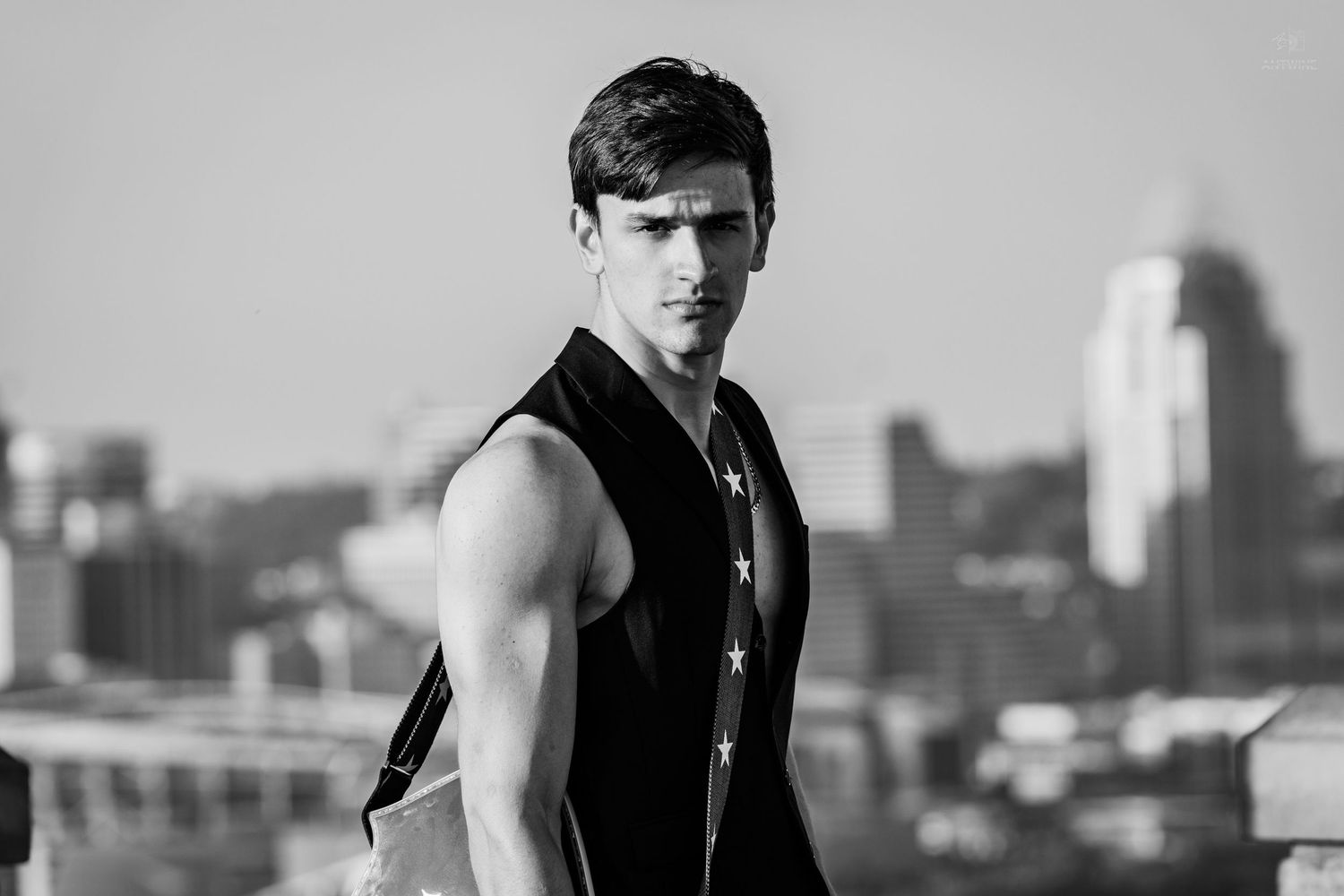 Muscular young man in sleeveless vest posing on rooftop with urban skyline in black and white.