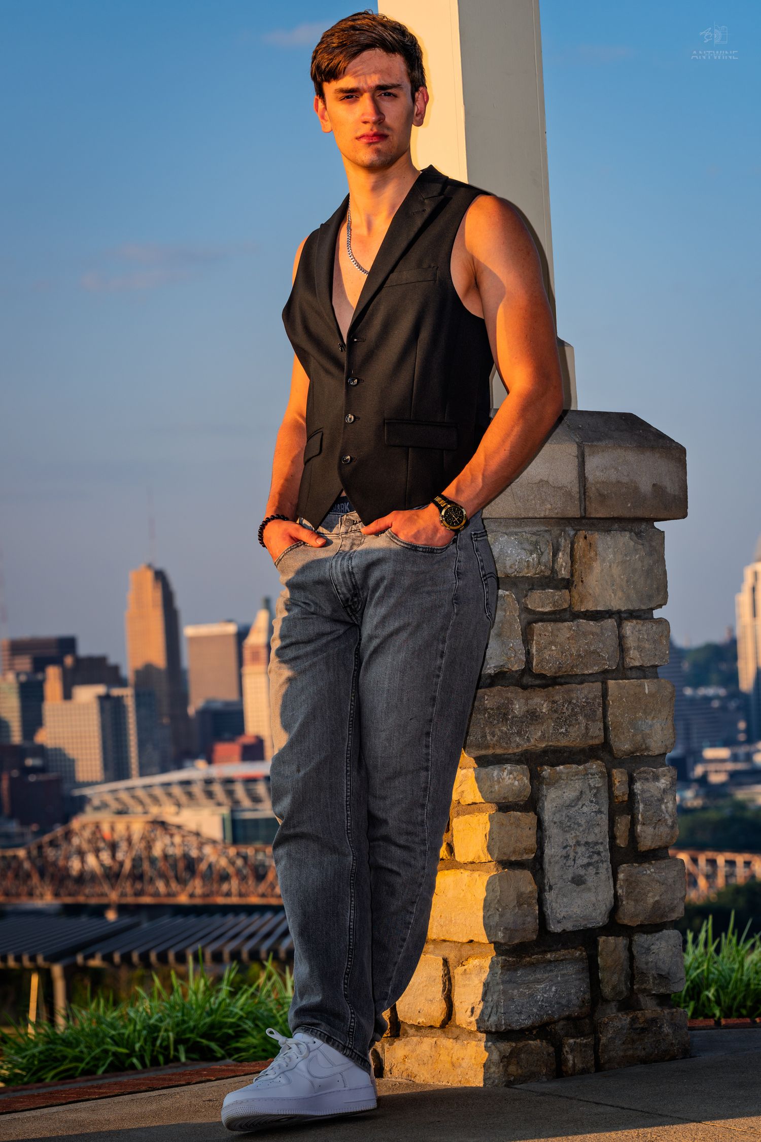 Young man in black vest and gray jeans leaning against stone pillar with city skyline at golden hour.