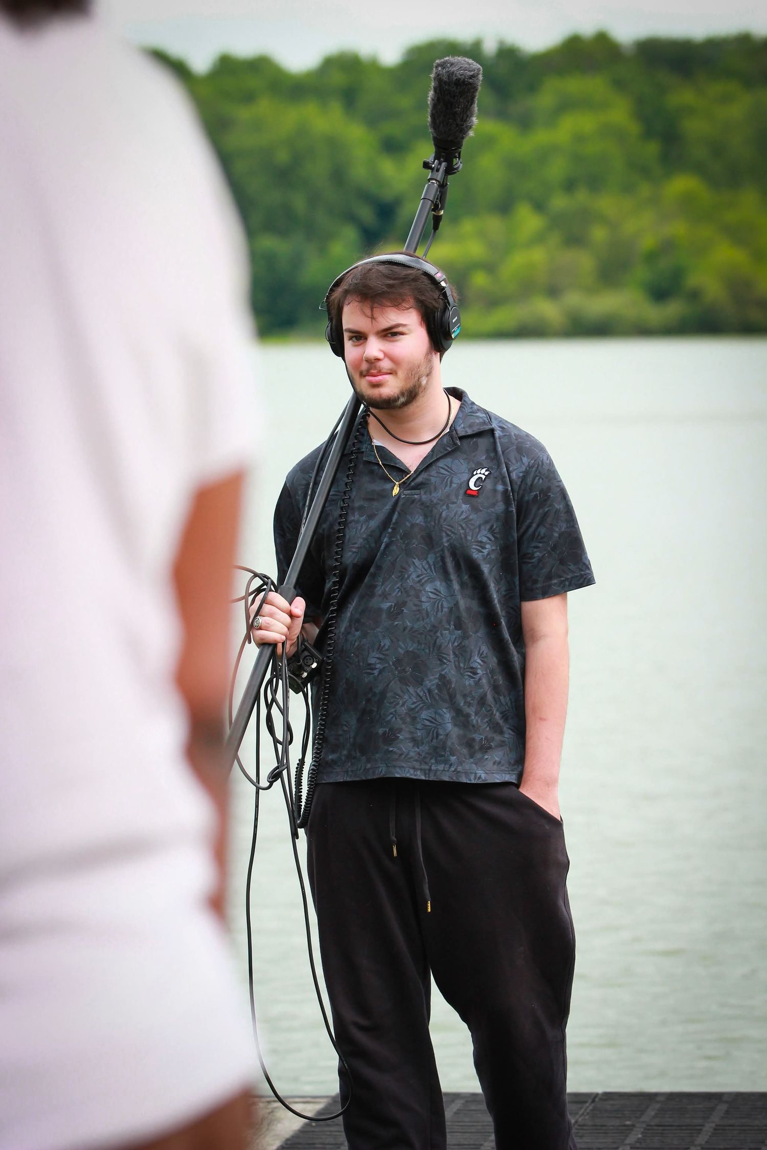 A young male sound technician wearing headphones holds a boom microphone near a lake outdoors.