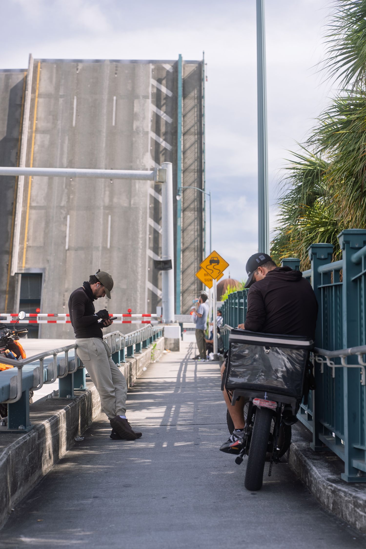 People waiting on a drawbridge sidewalk beside a tall concrete structure with a motorcycle nearby.