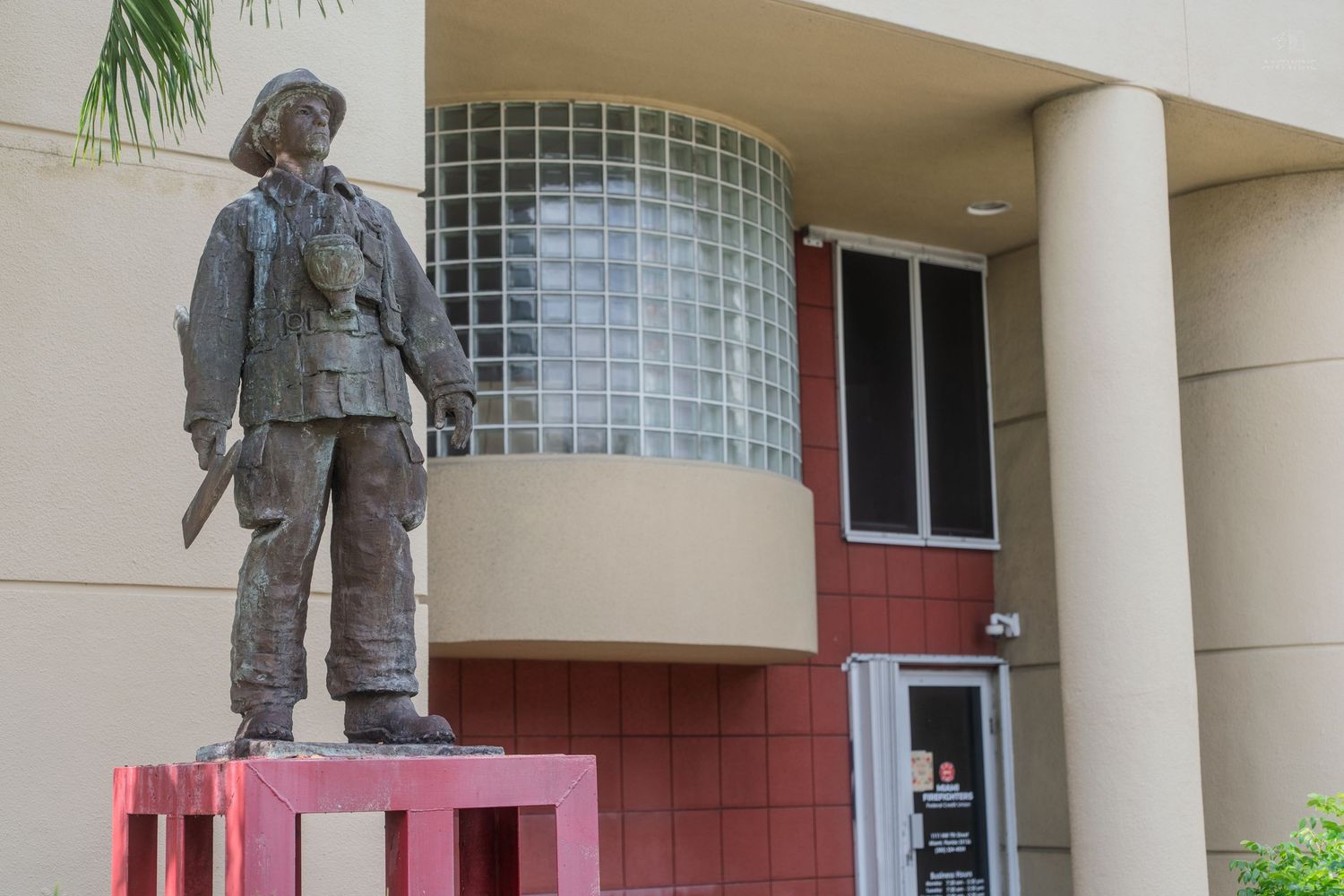 Bronze statue of a firefighter standing in front of a modern red and beige public building.