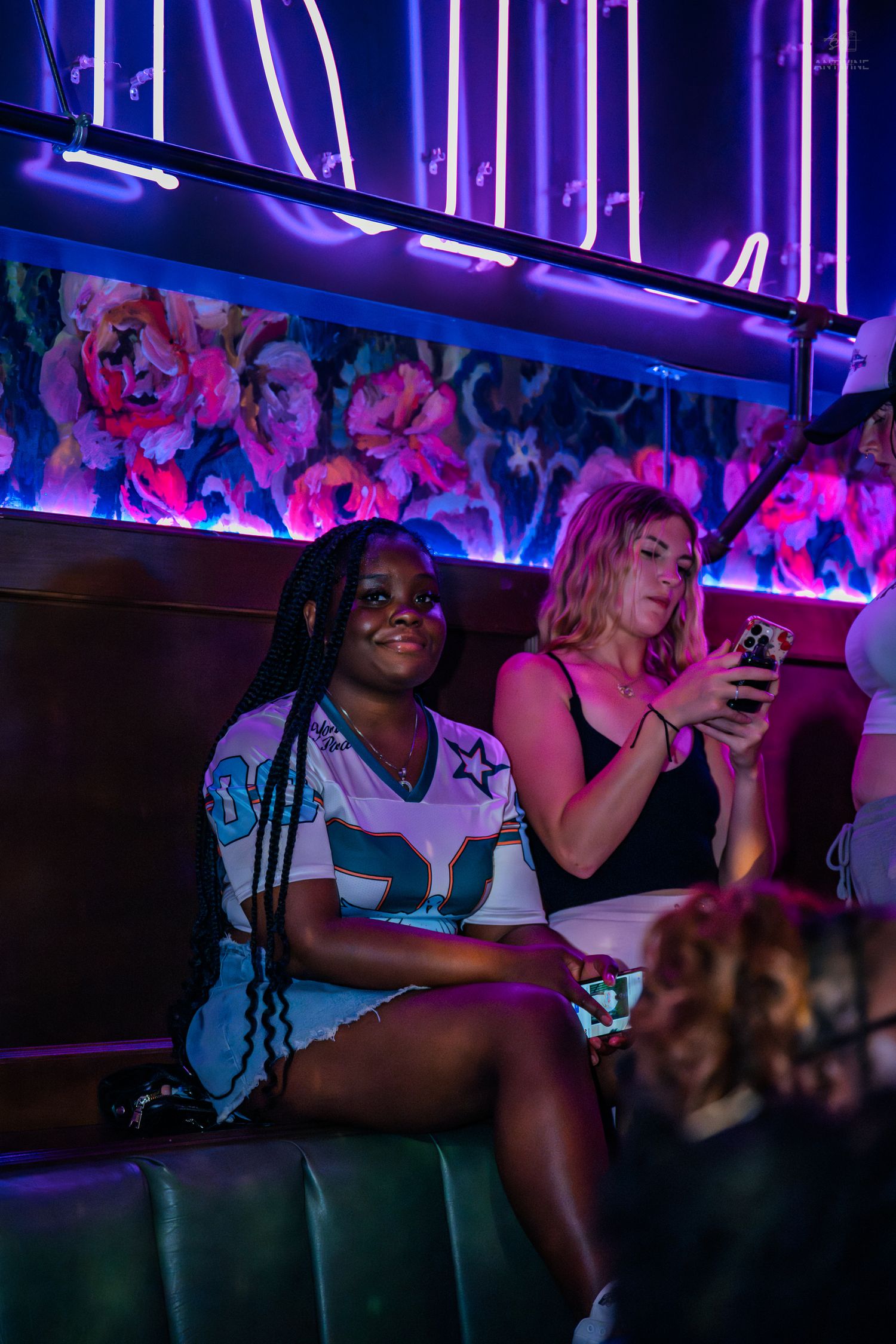 Two women sitting in a neon-lit nightclub booth with floral wall art and purple lighting.
