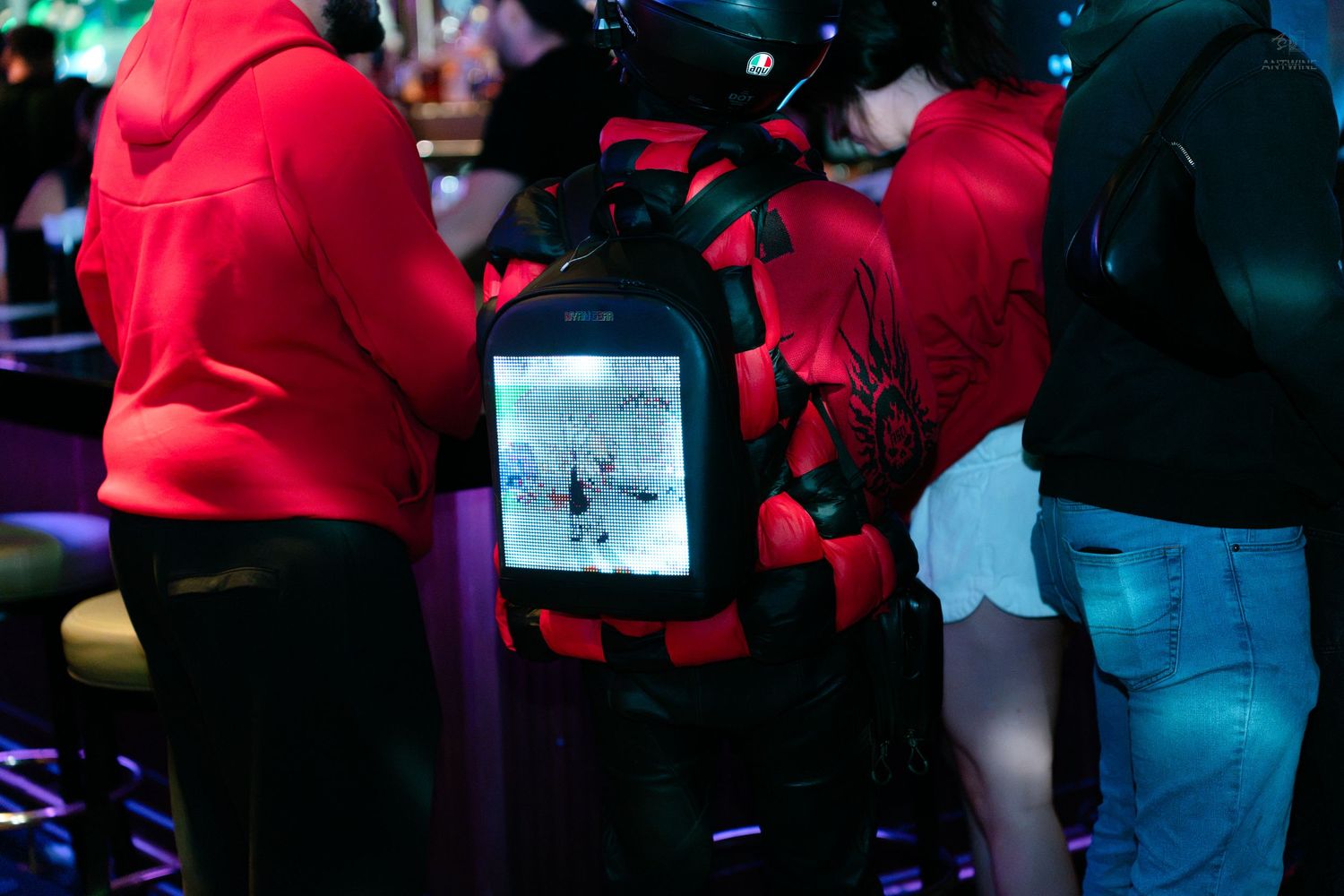 Person wearing a VR backpack gaming system with a lit screen display in a nightclub.
