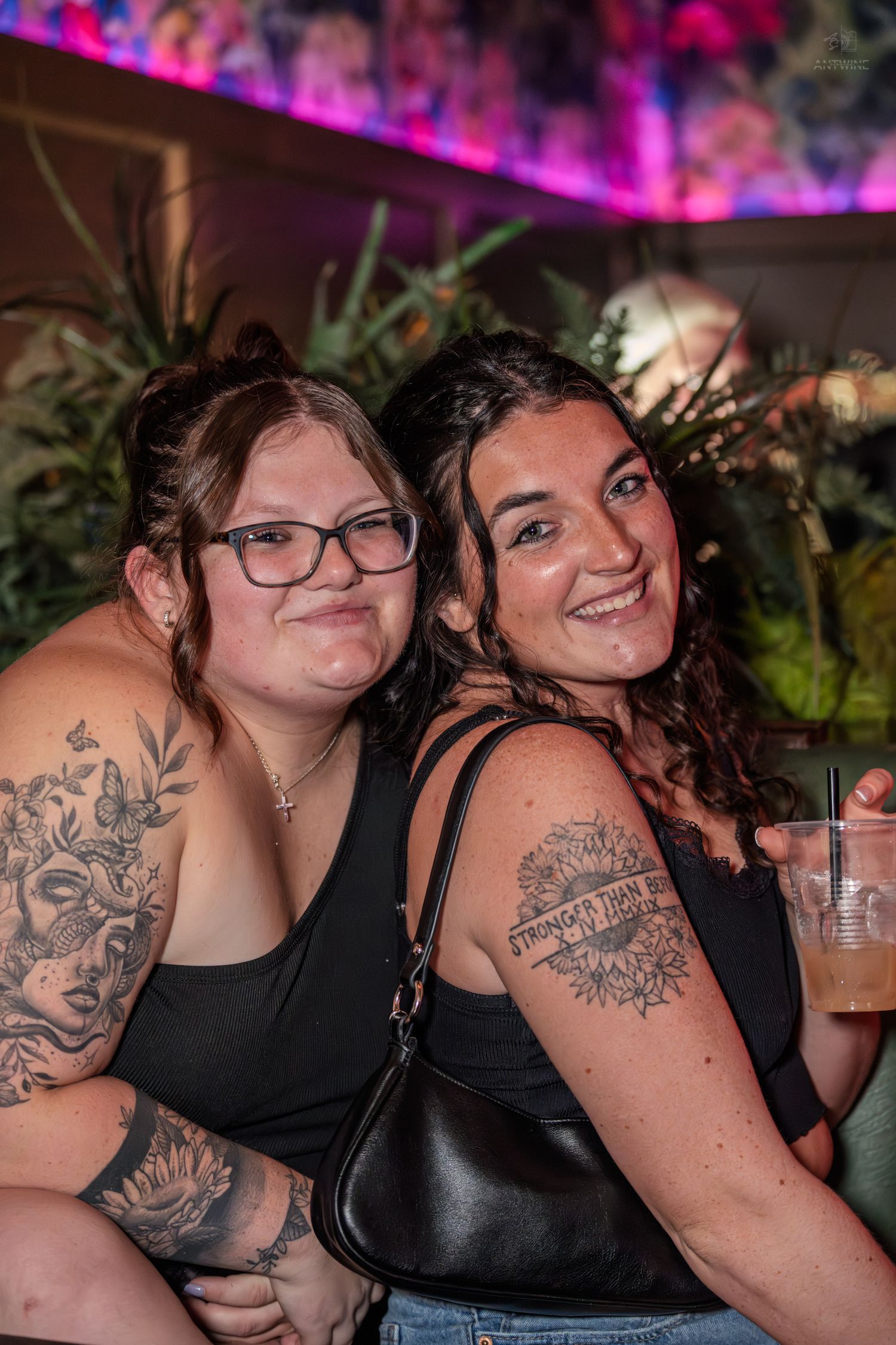 Two smiling tattooed women in black tops pose closely together in a vibrant nightclub lounge setting.