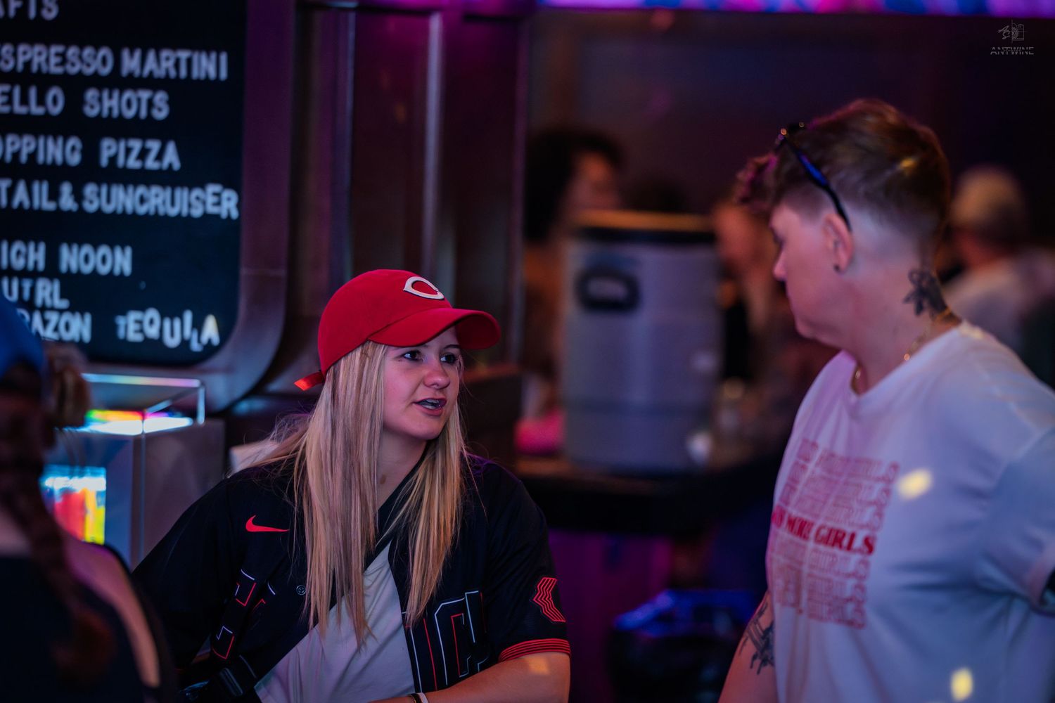 Blonde woman in Cincinnati Reds cap chatting at a vibrant nightclub bar.