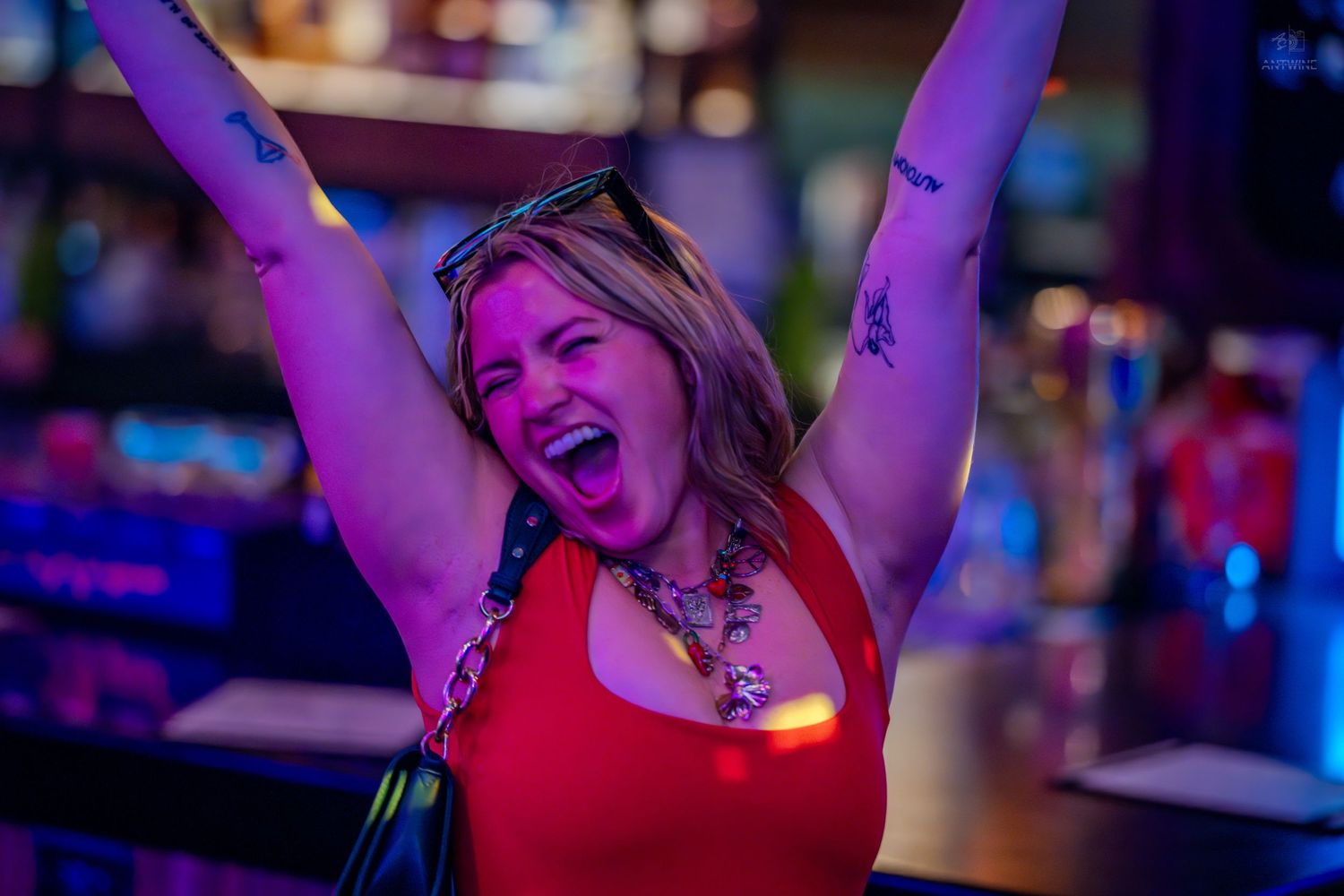 Excited woman in red top raises arms triumphantly at a lively bar celebration.