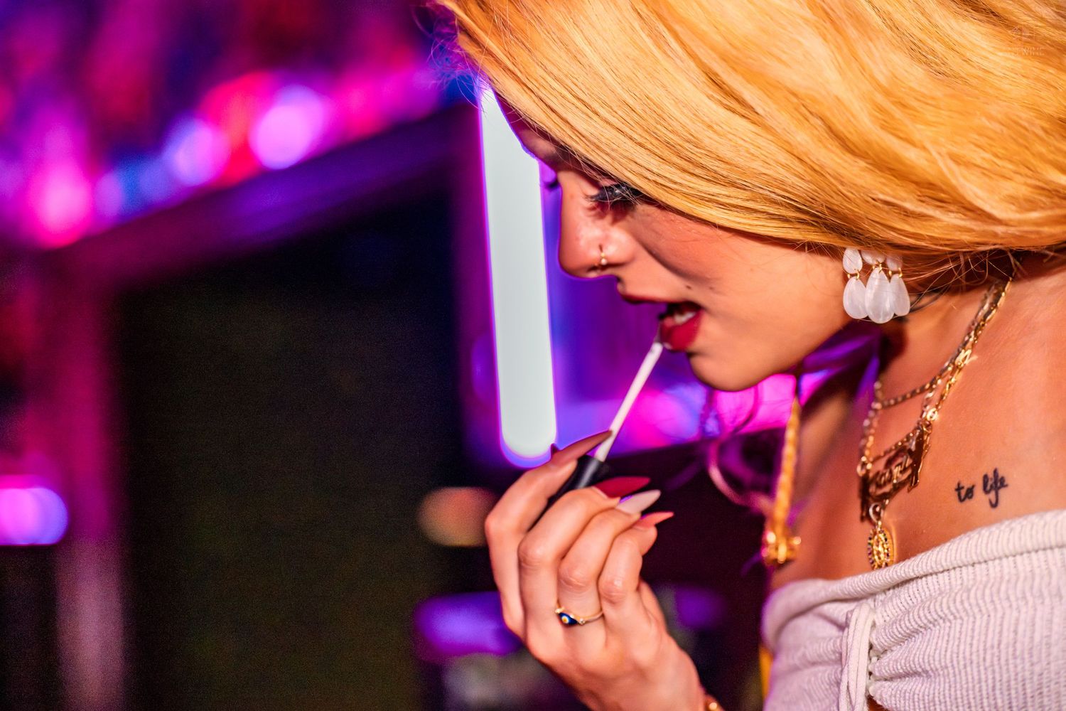 Close-up profile of blonde woman applying lip gloss under pink neon lights at a nightclub.