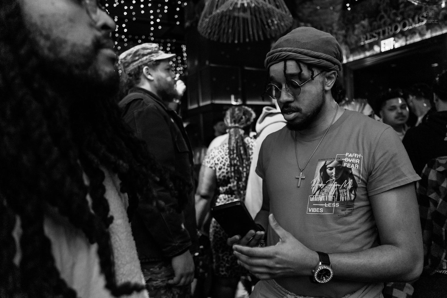 Man in cap and sunglasses checks phone at a crowded nightclub event in black and white.
