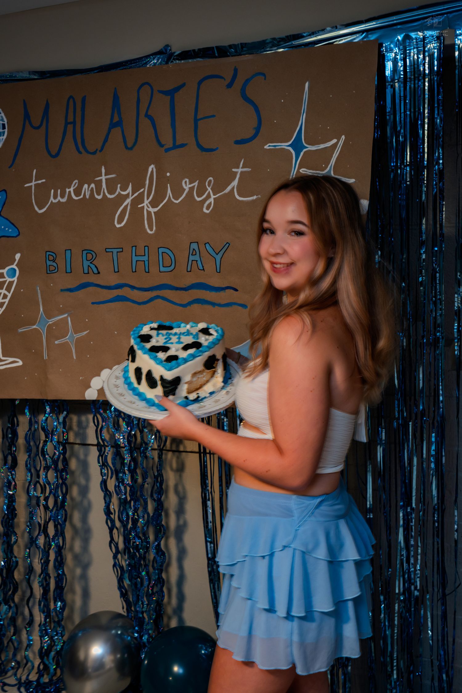 Smiling woman holding blue heart-shaped birthday cake sideways at Malarie's twenty-first birthday party with balloon decor.