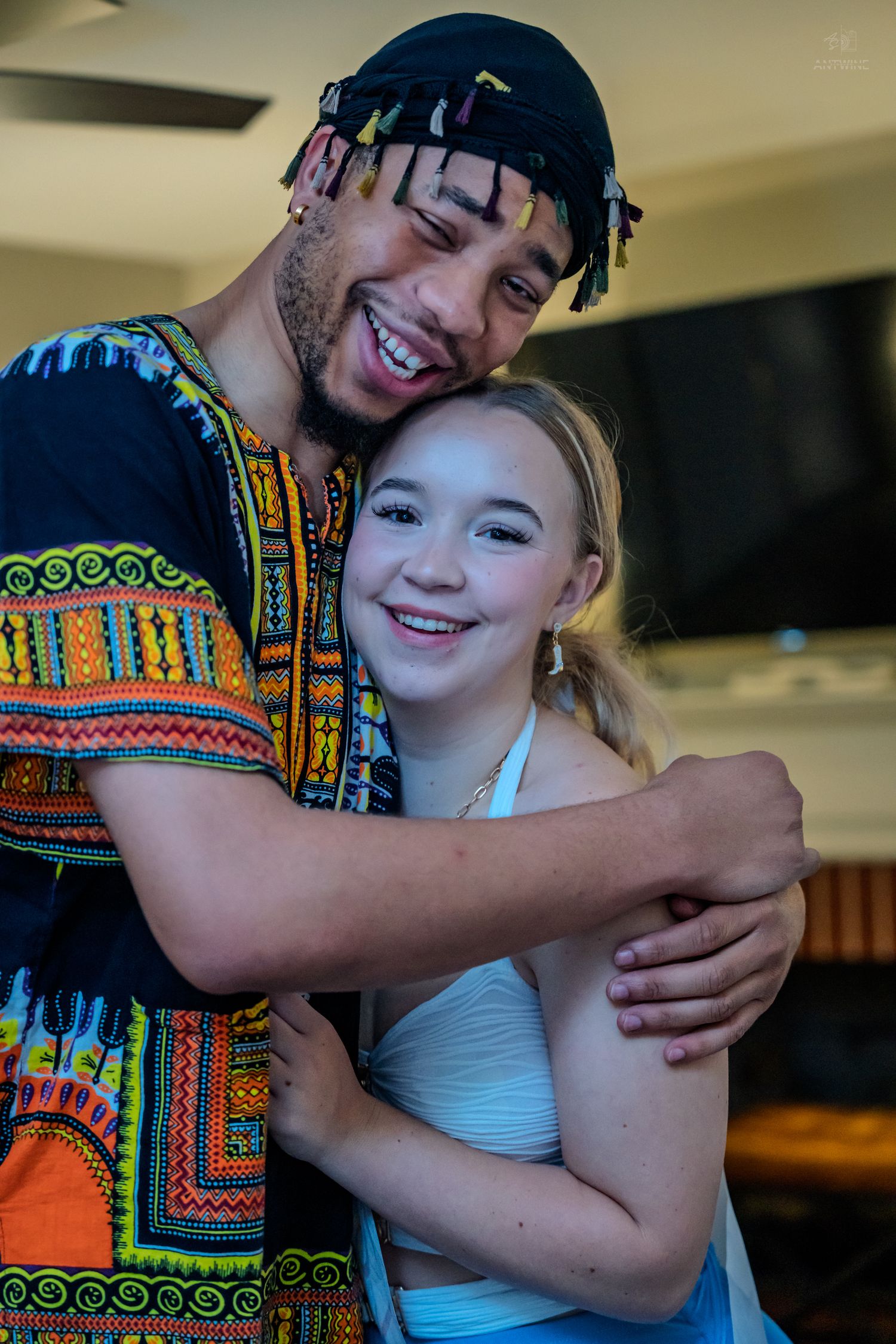 A smiling young man in a colorful dashiki hugs a happy young girl in a white top indoors.