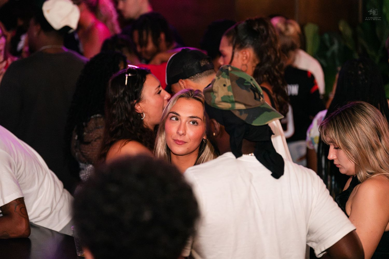 Crowd of young people mingling at an indoor nightclub party with pink lighting.