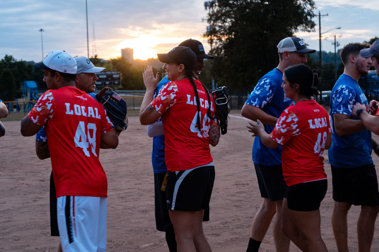 Players in red Local jerseys line up on a softball field at sunset during a charity game.