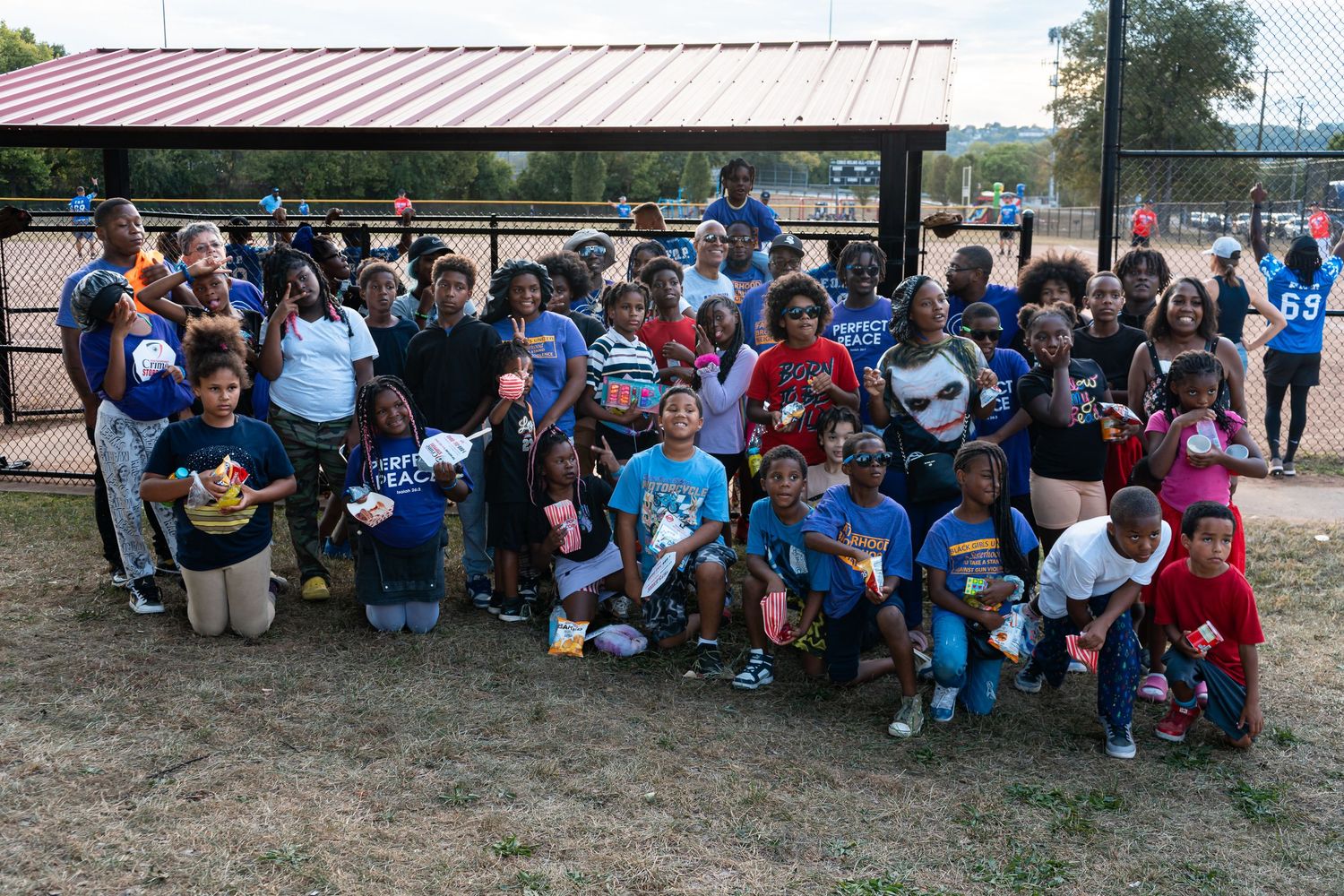 Large group of diverse children and adults pose together for a photo at an outdoor baseball field event.