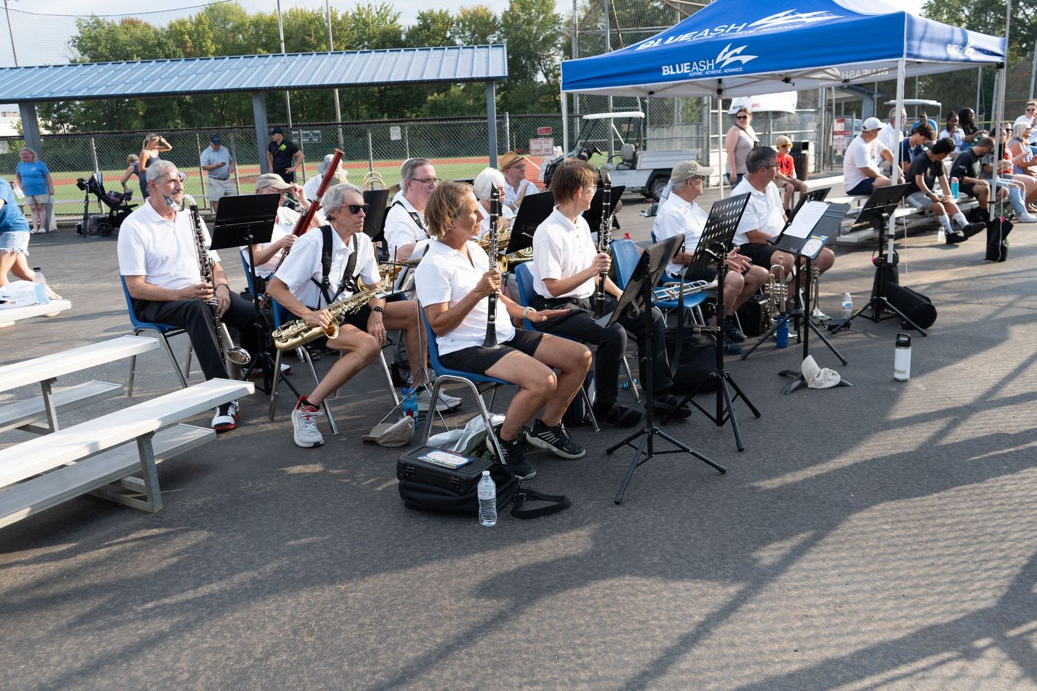 Outdoor band performance with musicians in white shirts playing instruments on bleachers under a blue tent on a sunny day.