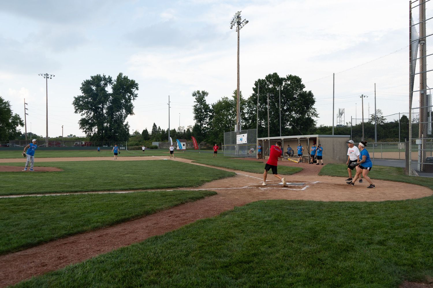Players compete in a recreational softball game on a green field at dusk, with teams in red and blue jerseys.