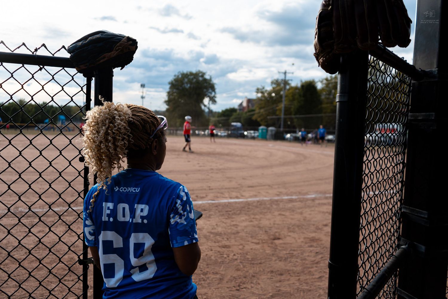 A child in a blue FOP 69 jersey watches a softball game through a chain-link fence on a cloudy day.