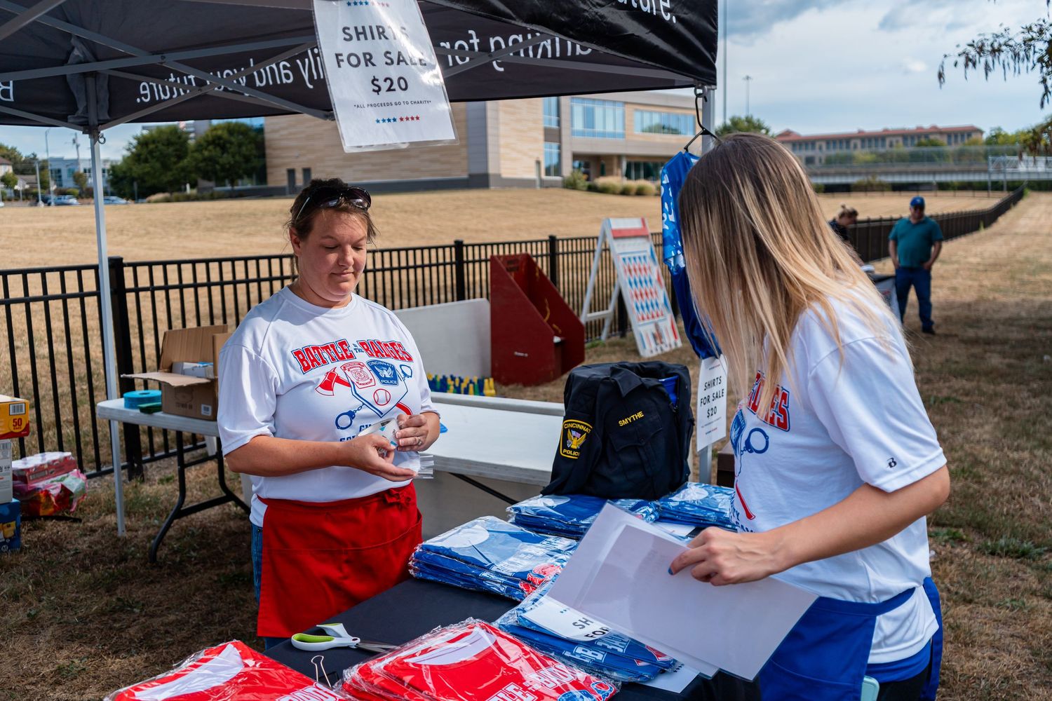 Two women at an outdoor merchandise booth selling blue T-shirts at a sunny community event.