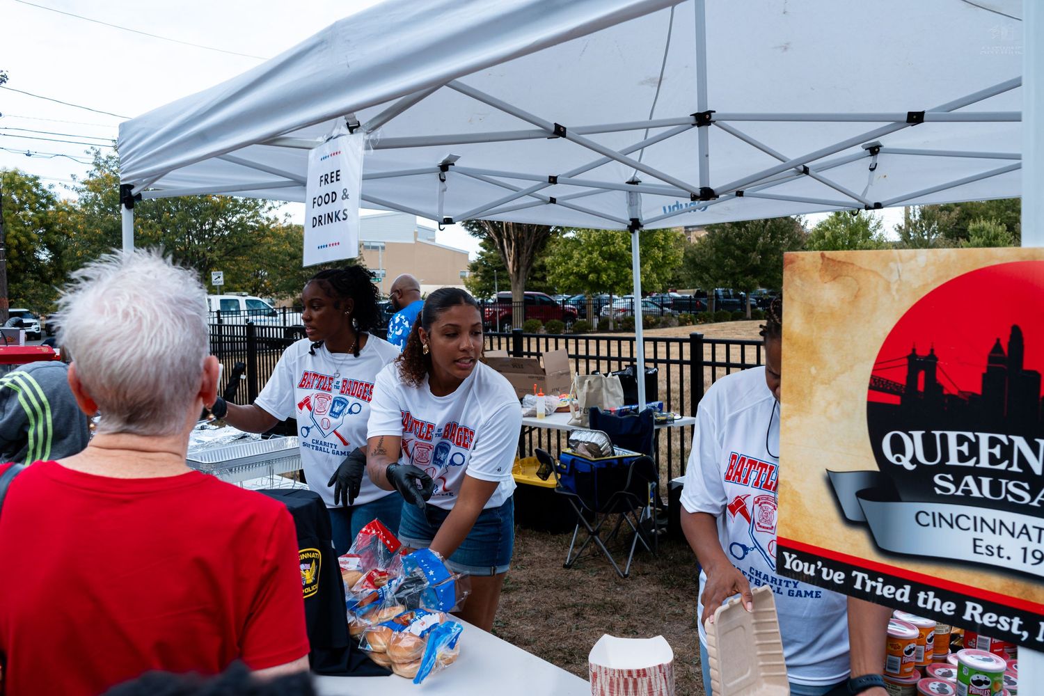Volunteers serve food at a Queen City Sausage booth under a white tent at an outdoor community event.