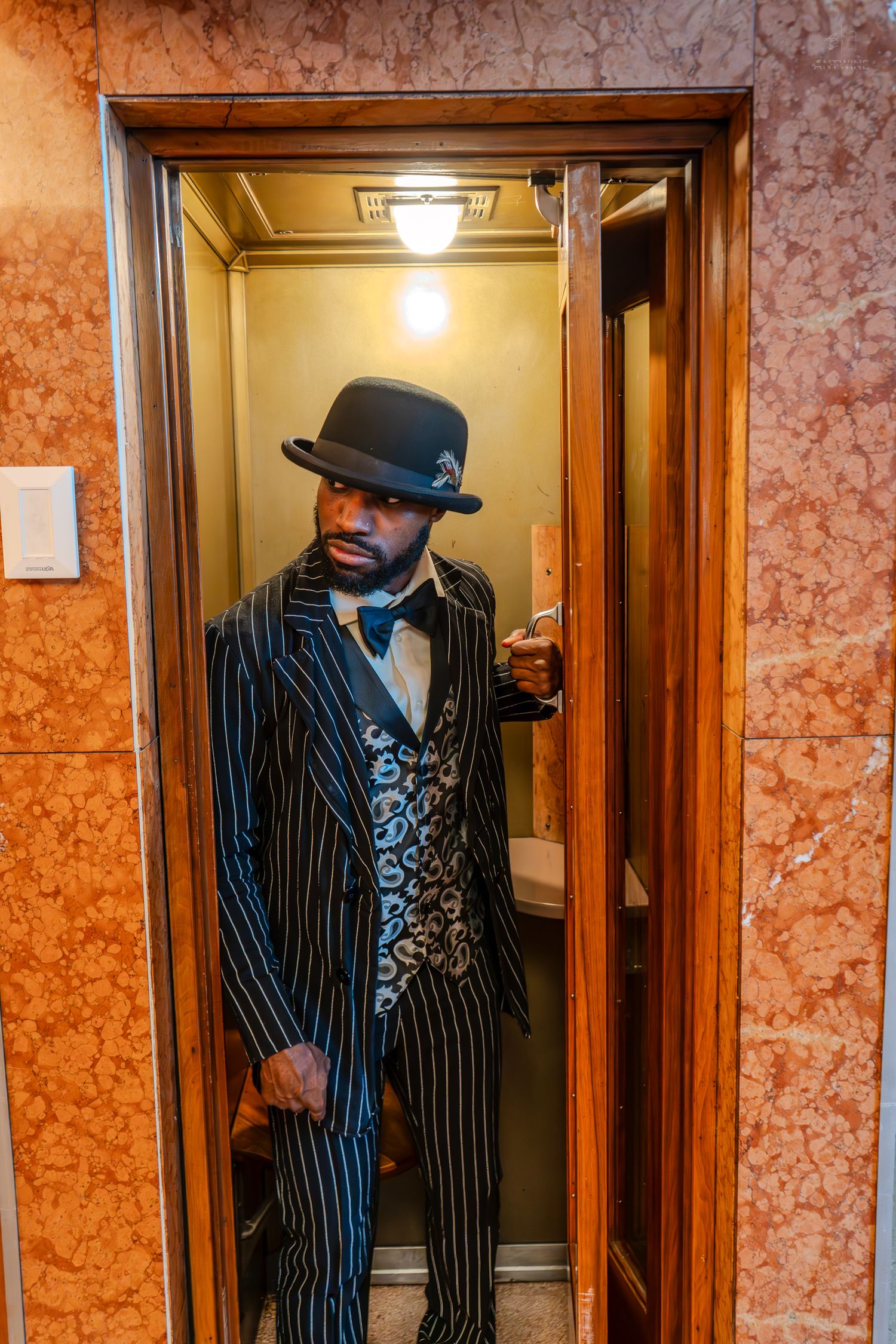 Man in pinstripe suit, bow tie, and bowler hat posing in an elegant elevator with marble walls.