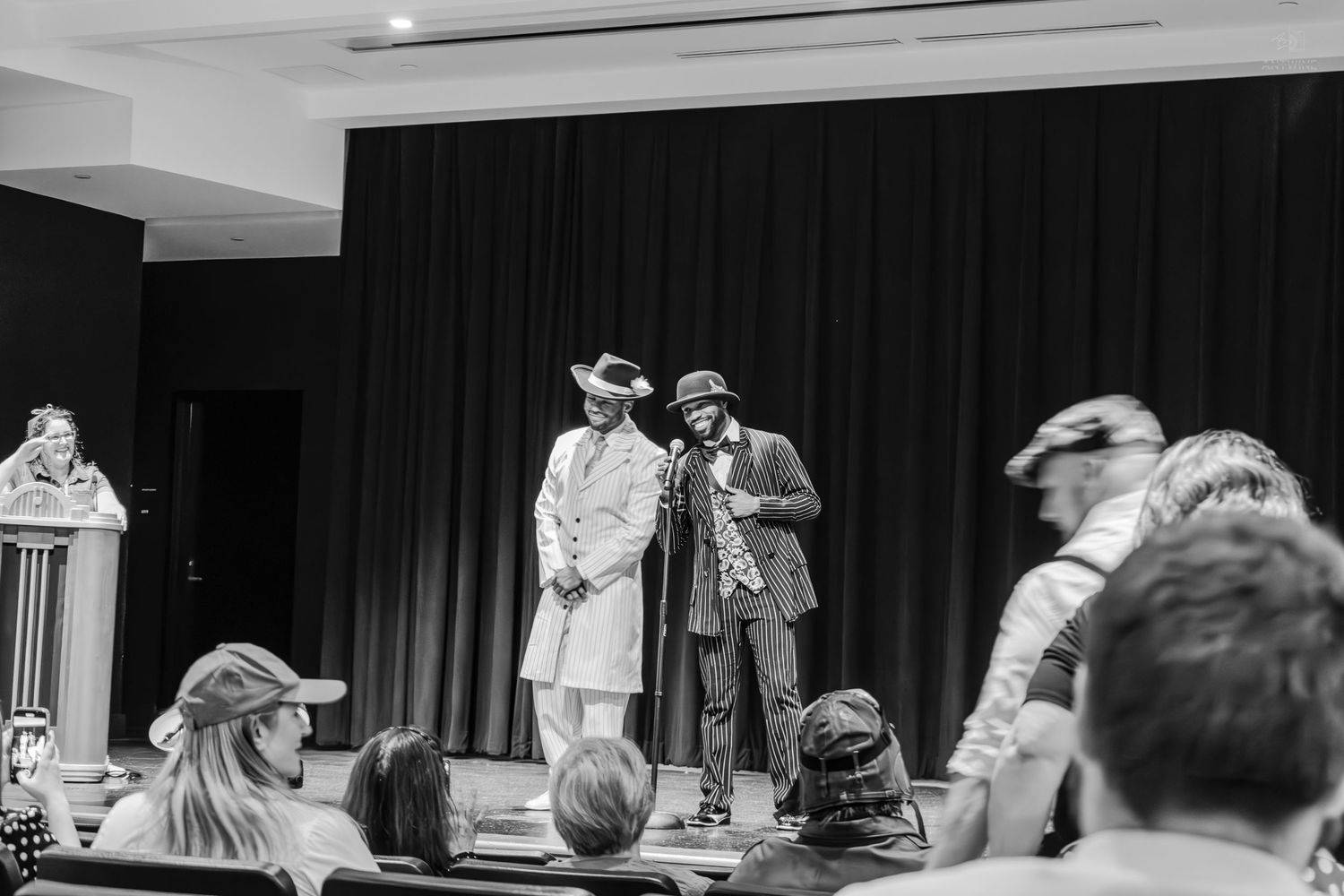 Two performers in costume on stage at a live theater event, with audience members seated in the foreground.