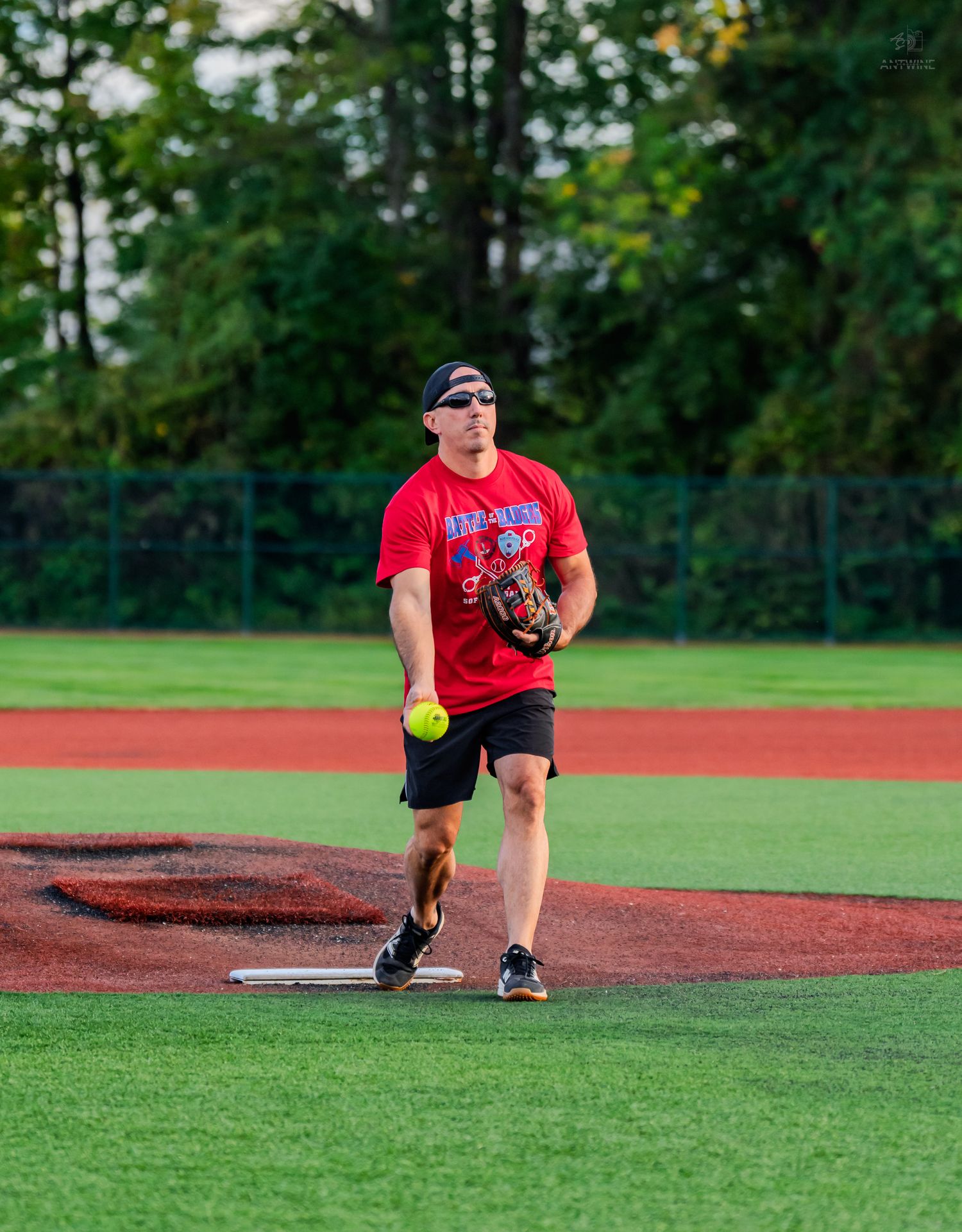 Male softball player in red shirt pitching a yellow ball on a green field at dusk.
