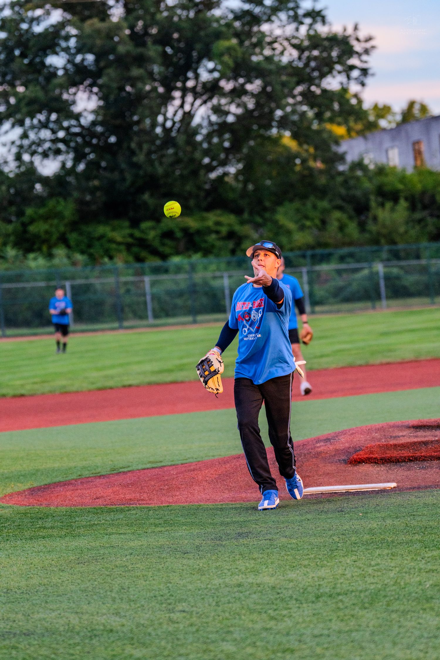 Female softball pitcher releasing yellow ball on pitcher mound during evening game.