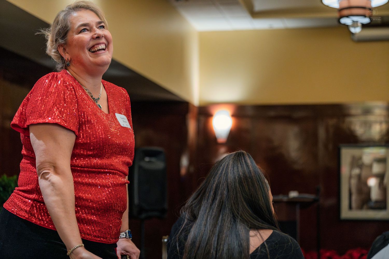 Smiling woman in red sequin top stands confidently at a formal indoor gathering.