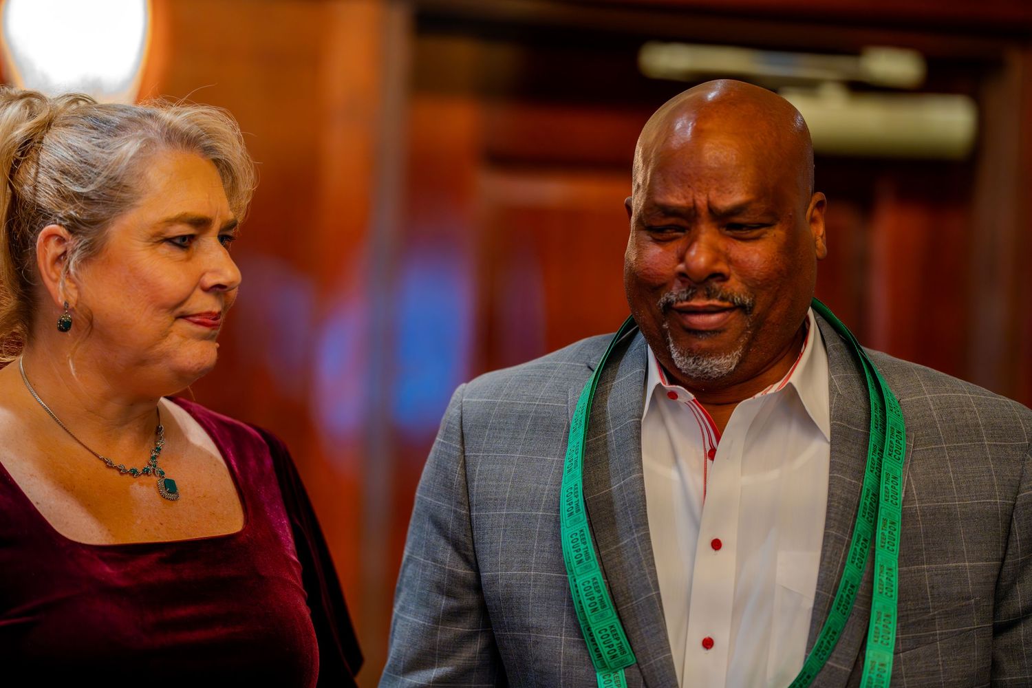 A man in a gray suit with green sash stands beside a woman in burgundy velvet dress at a formal event.