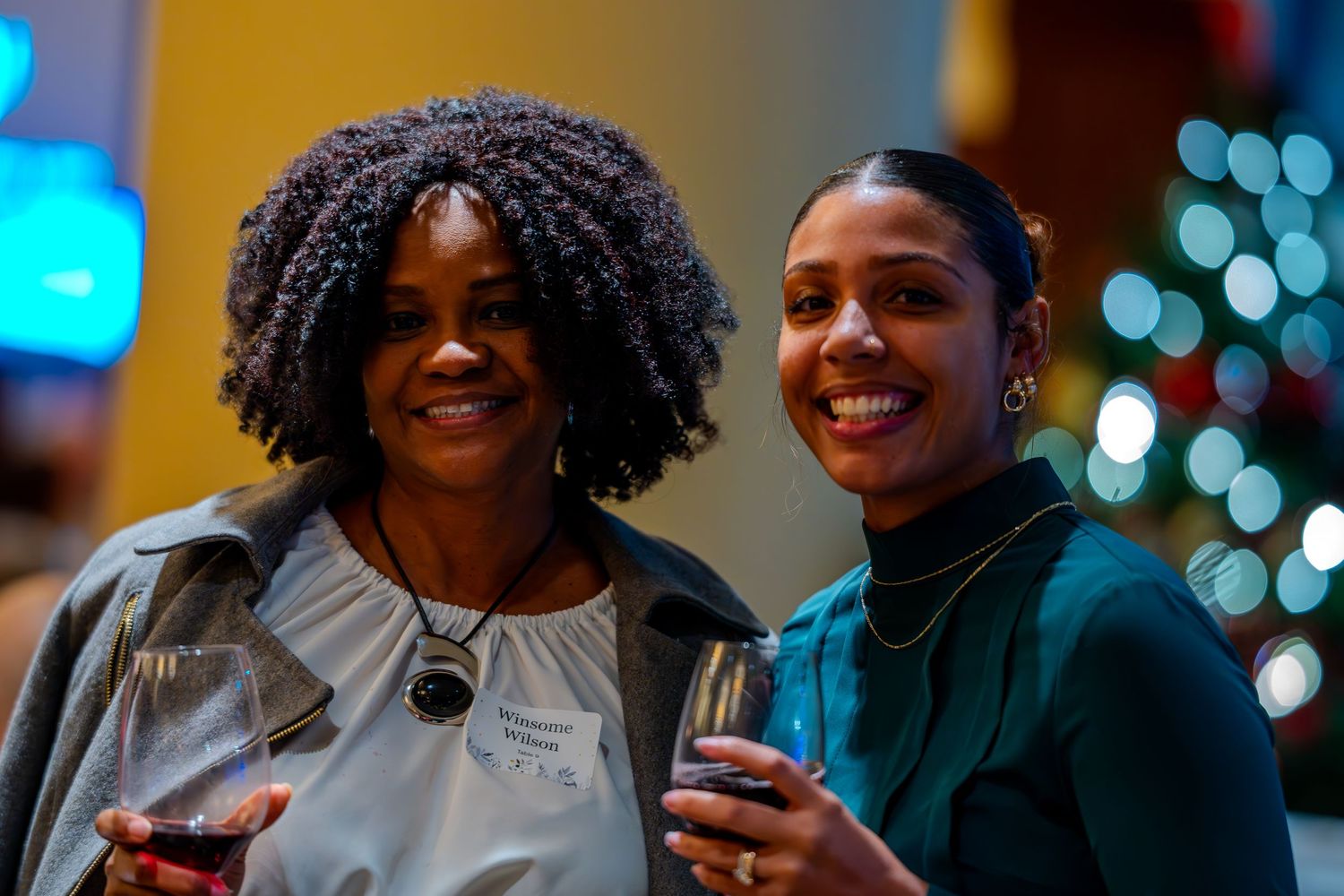 Two smiling women holding wine glasses pose together at a festive holiday party near a Christmas tree.