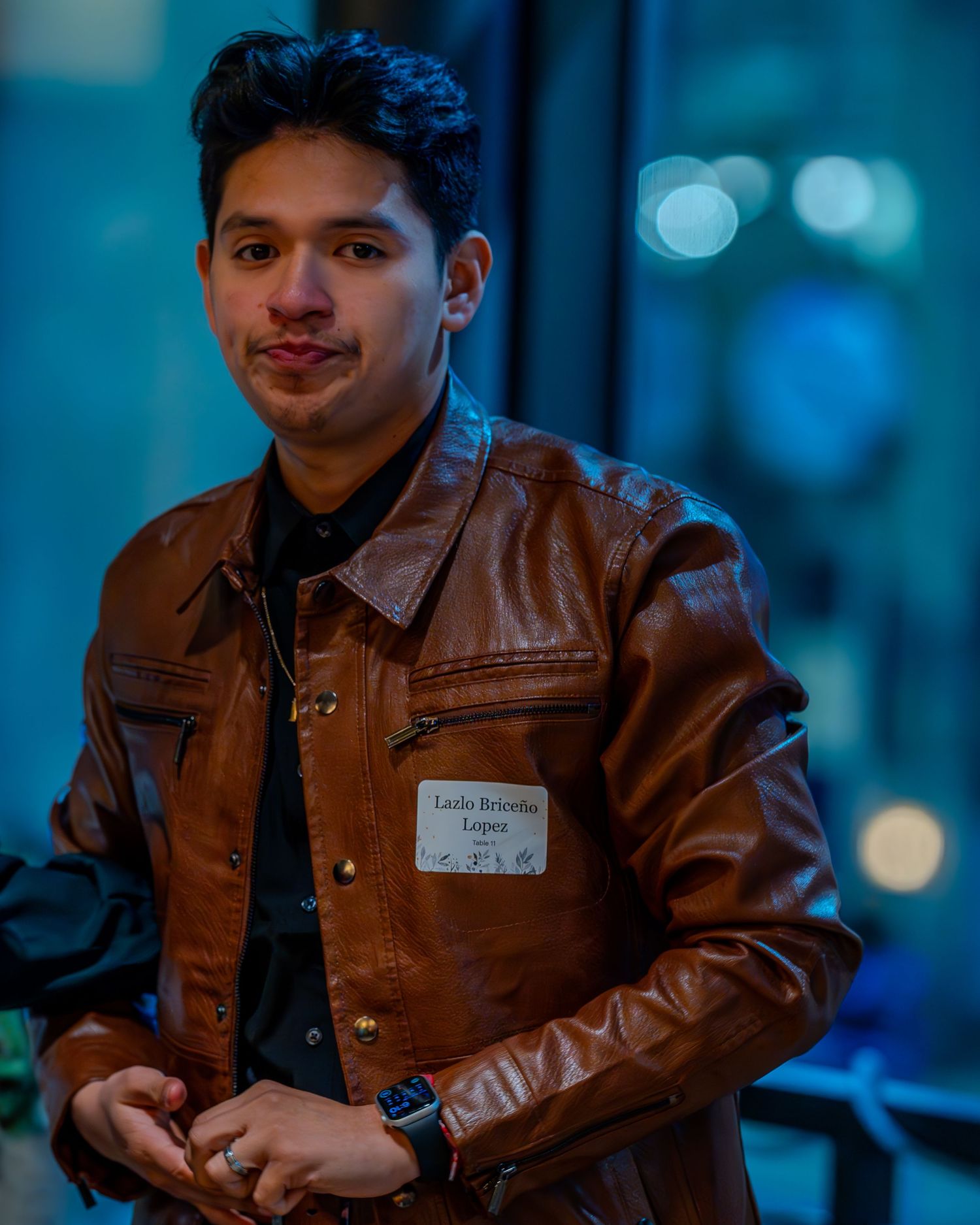 Young man in brown leather jacket with name tag smiling at indoor event with blue bokeh background.