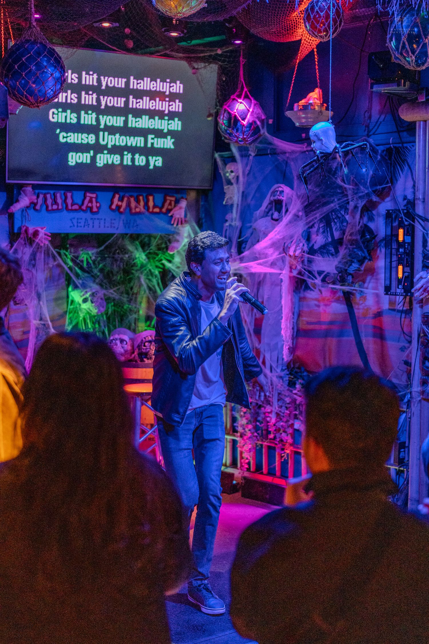 Man singing karaoke at Halloween-decorated Hula Hula bar in Seattle with colorful neon lights.
