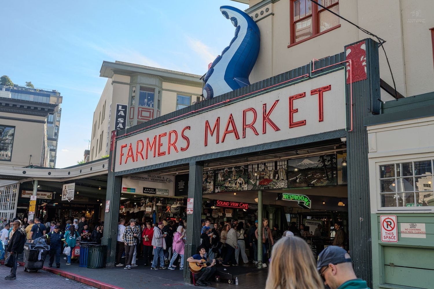 Crowds gather outside the iconic Pike Place Farmers Market sign with a large blue fish sculpture in Seattle, WA.