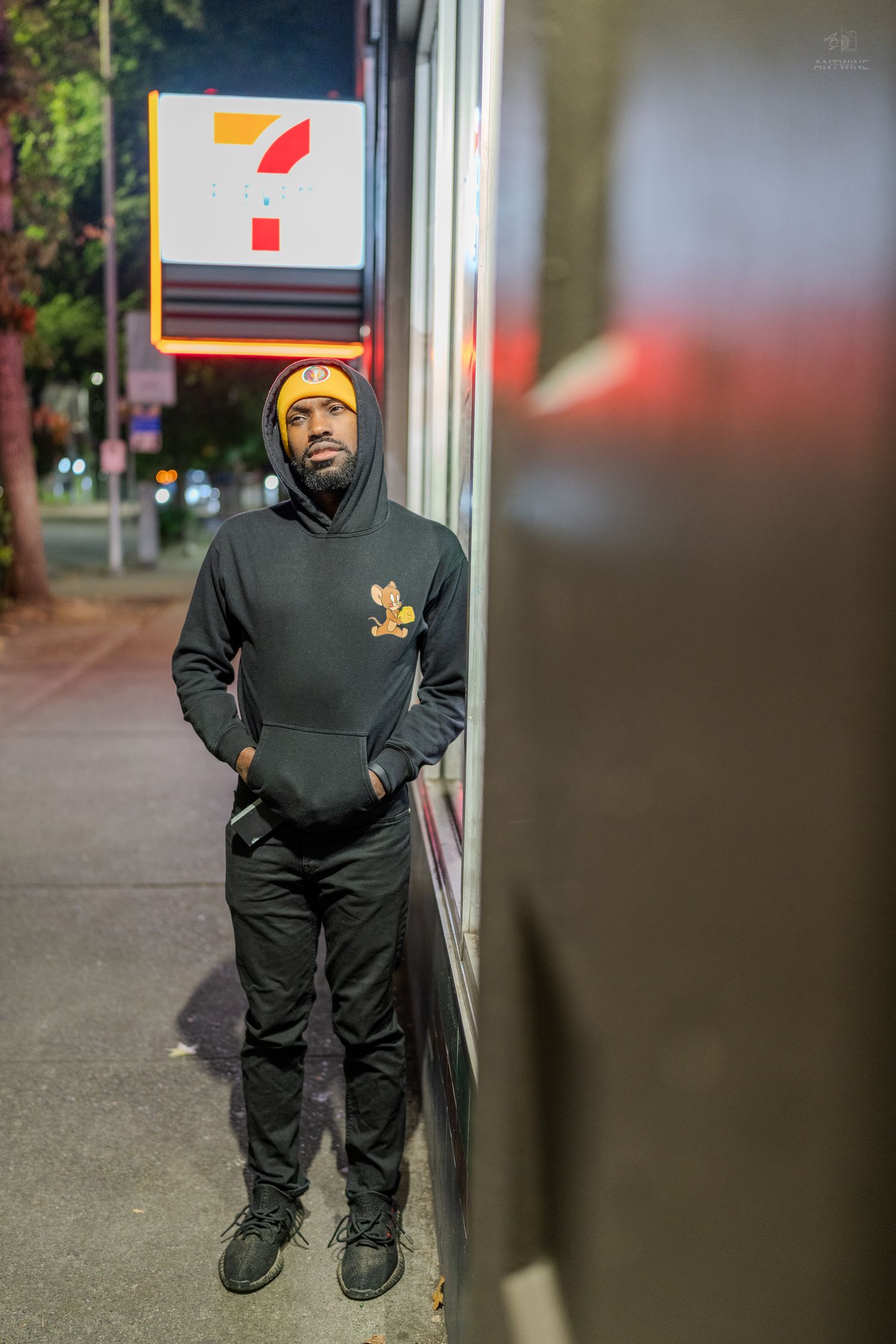 Man in black hoodie and yellow beanie standing outside a 7-Eleven store at night.