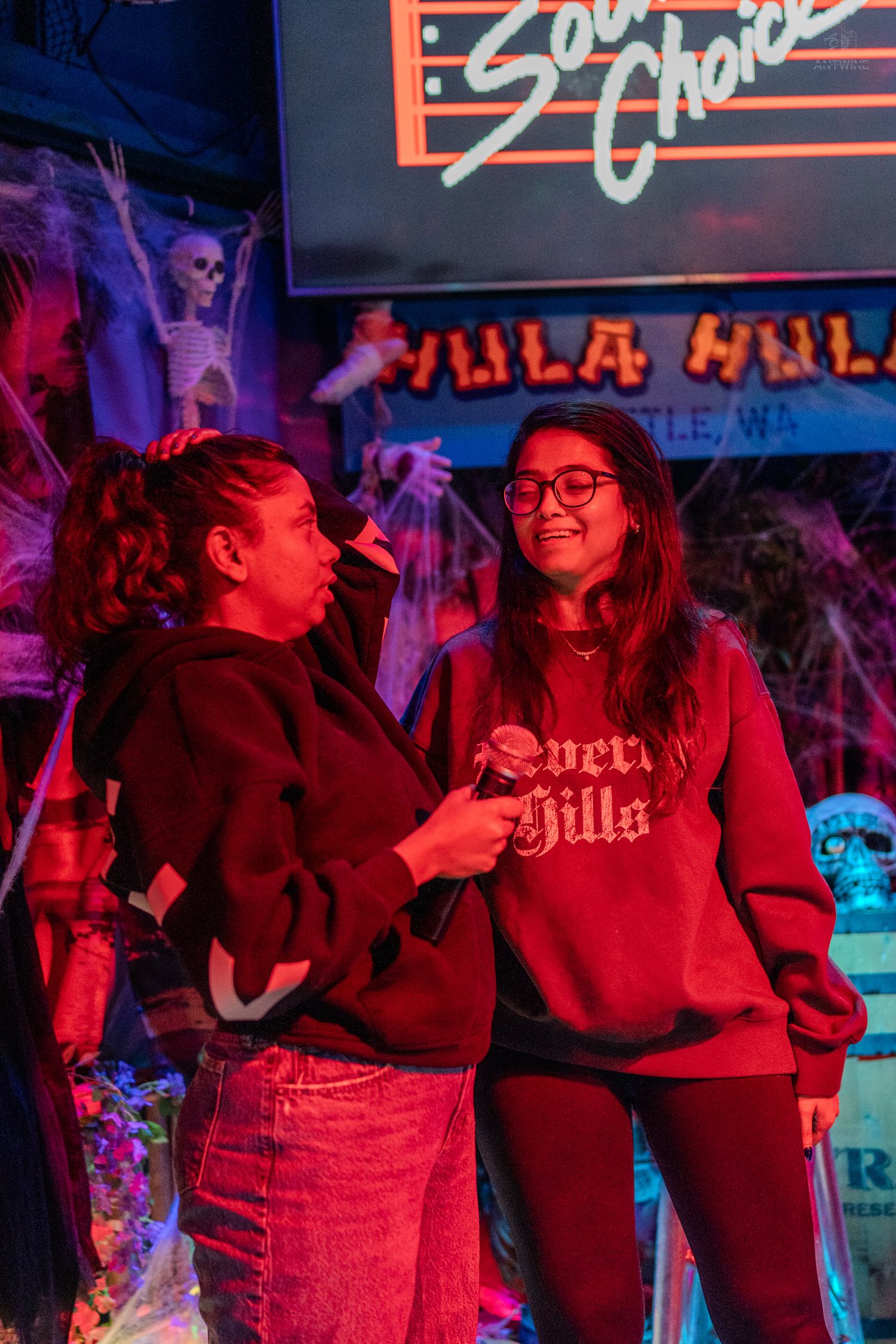 Two women singing karaoke at Hula Hula bar in Seattle, WA, decorated with Halloween skeletons and purple neon lights.
