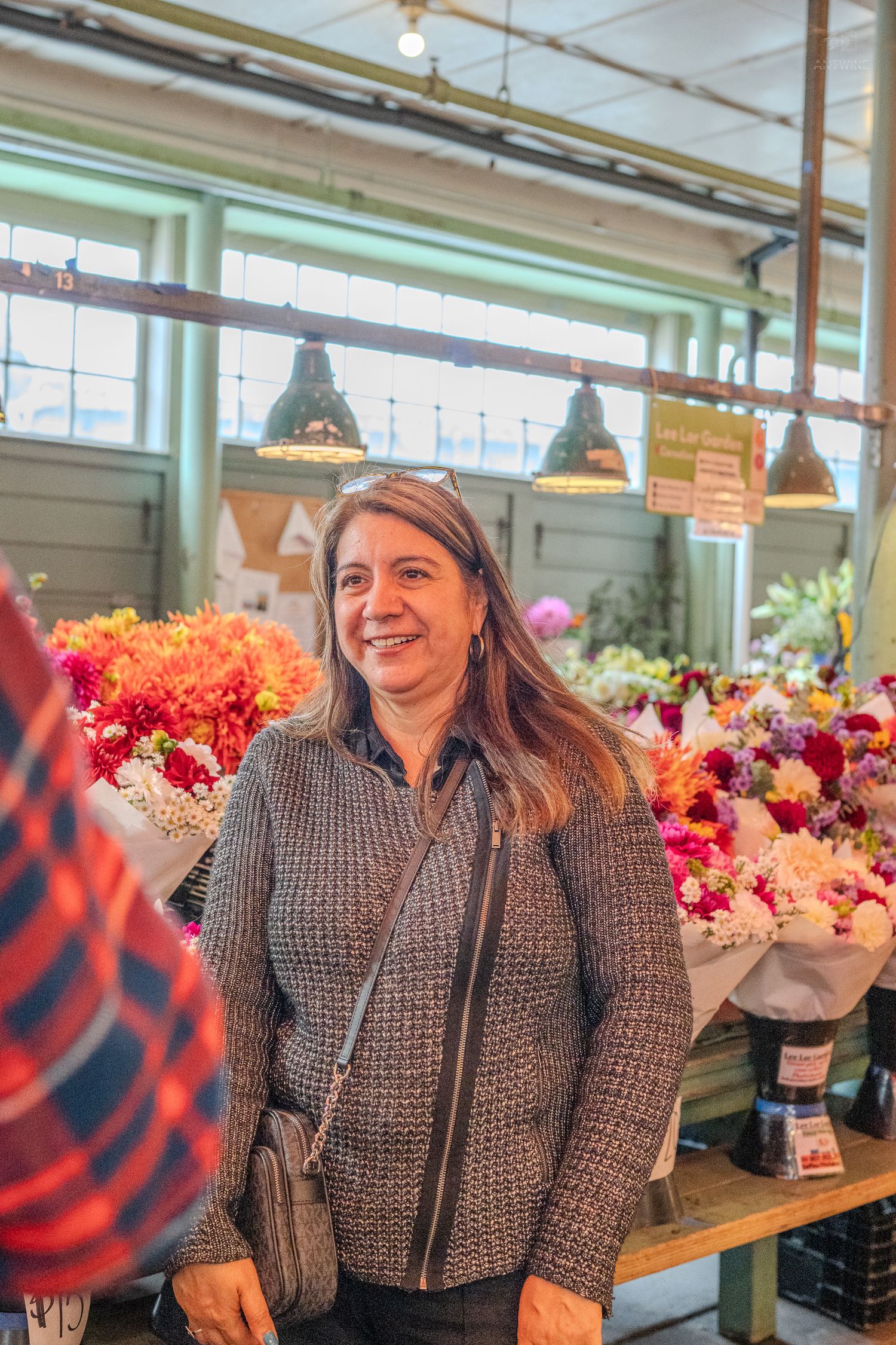 Smiling woman in gray jacket standing in front of colorful flower bouquets at Pike Place Market.