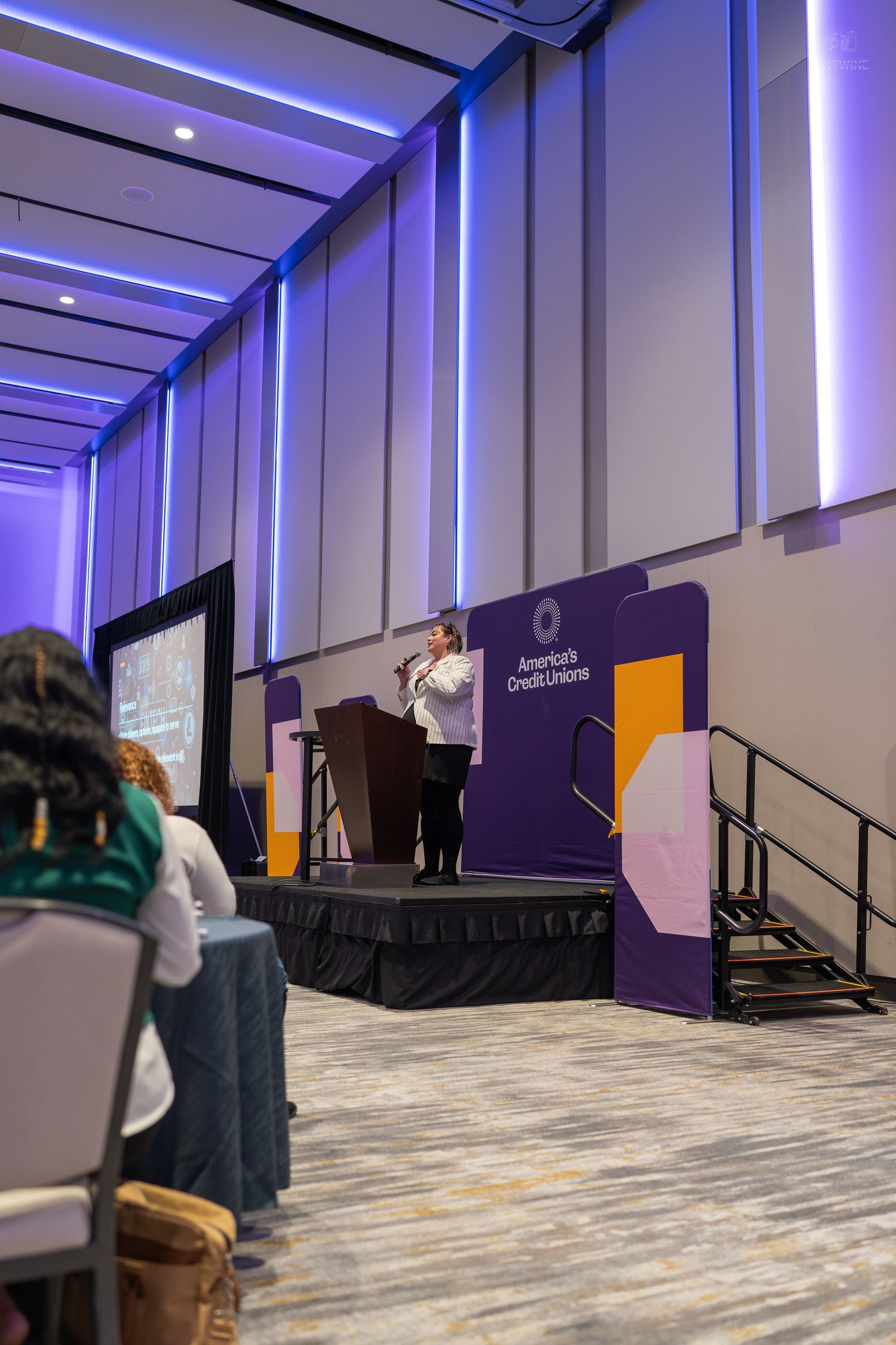 Speaker presents at podium on stage at America's Credit Unions event in a conference hall with blue accent lighting.