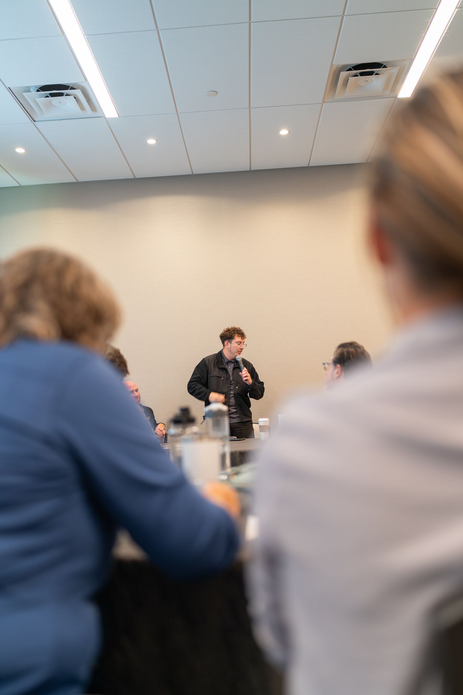 A speaker presents to a small group of professionals seated at tables in a modern conference room.