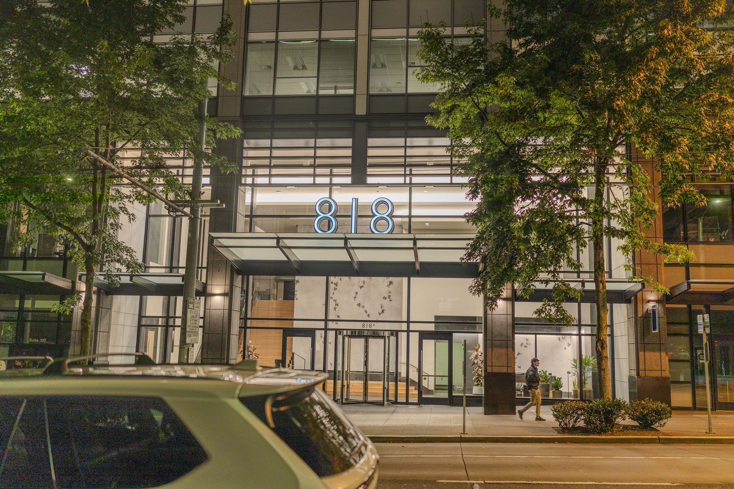 Modern glass office building at 818 illuminated at night with lush green trees and parked cars on an urban street.