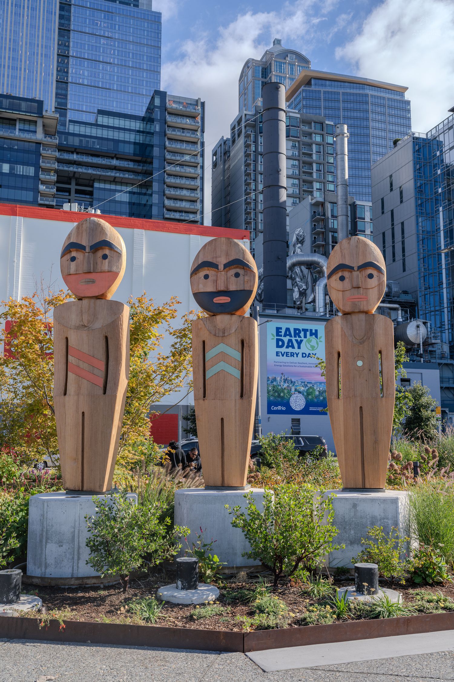 Three carved wooden Indigenous totem figures displayed outdoors with Seattle glass skyscrapers and Earth Day sign behind.