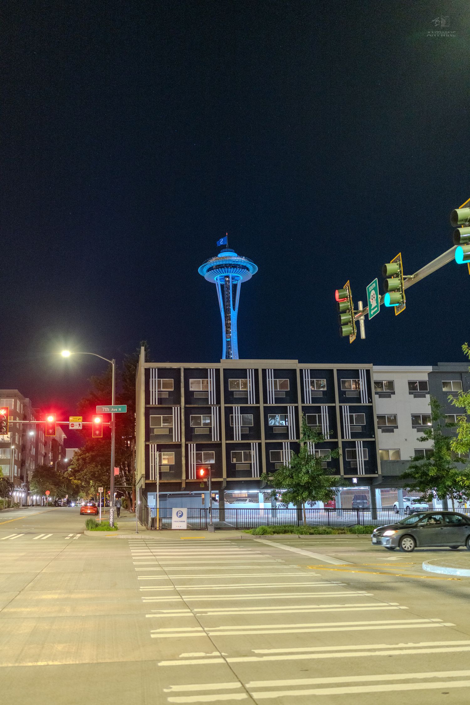 Seattle Space Needle glowing blue at night, viewed from a city intersection with traffic lights and modern apartment buildings.