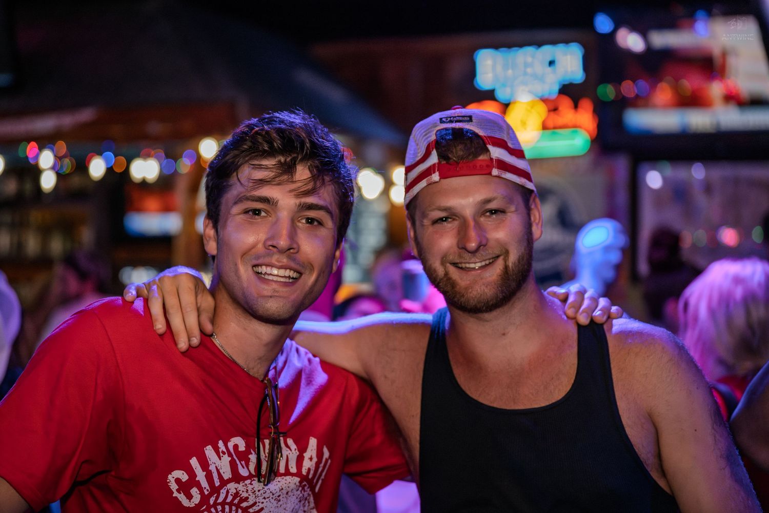 Two smiling men pose together at a lively bar with colorful neon lights in the background.