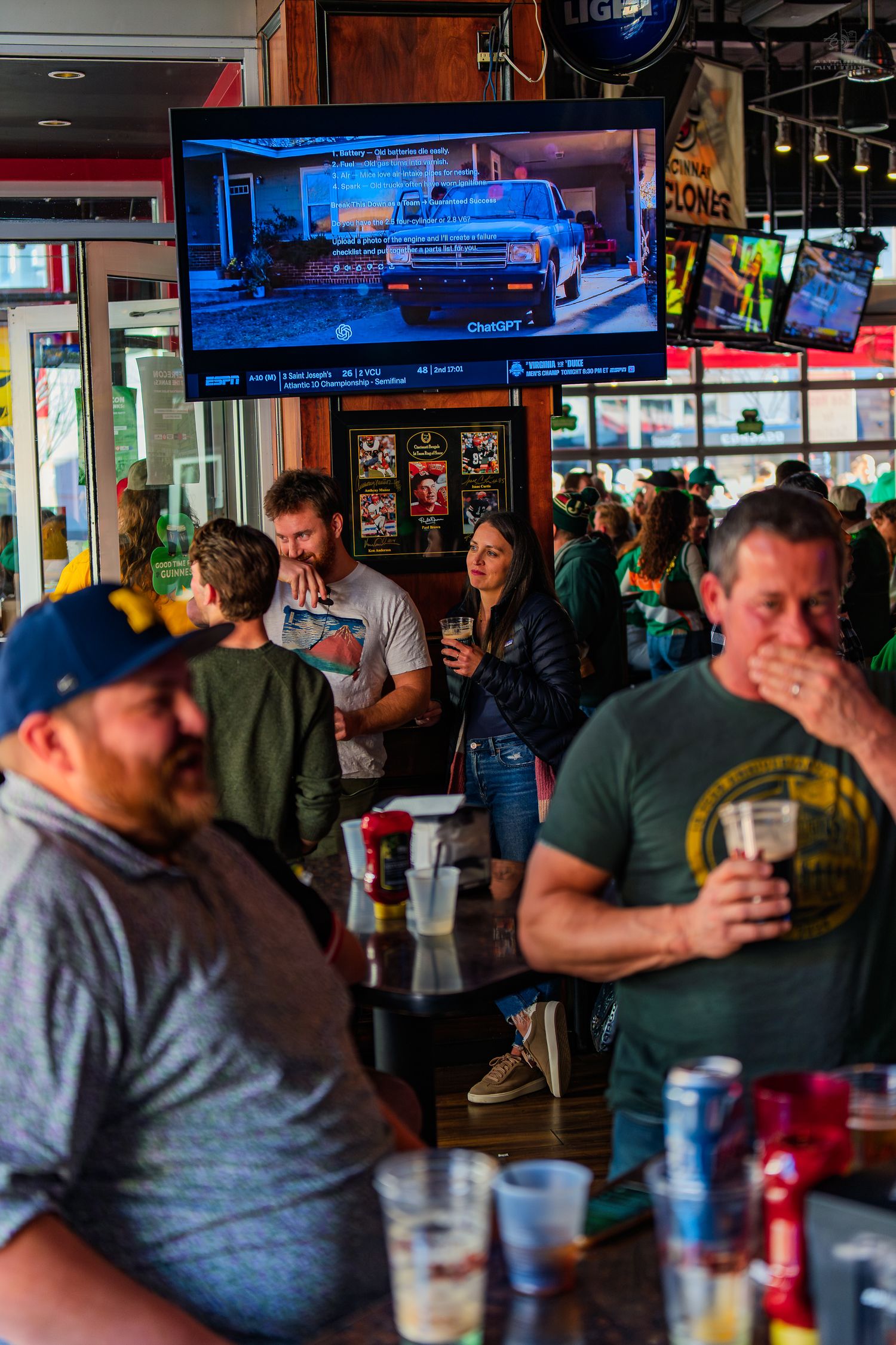 Busy bar scene with patrons drinking and socializing, TV displaying a truck show, green decorations suggesting St. Patrick's Day.