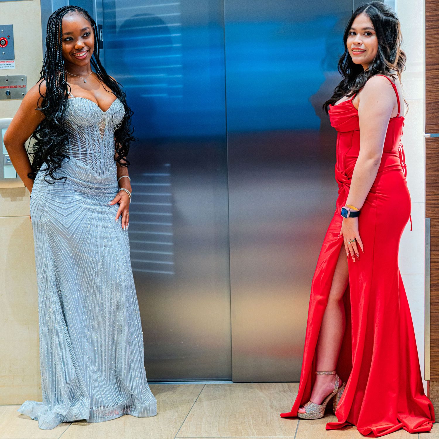 Two women in formal gowns, one silver and one red, posing near elevator.