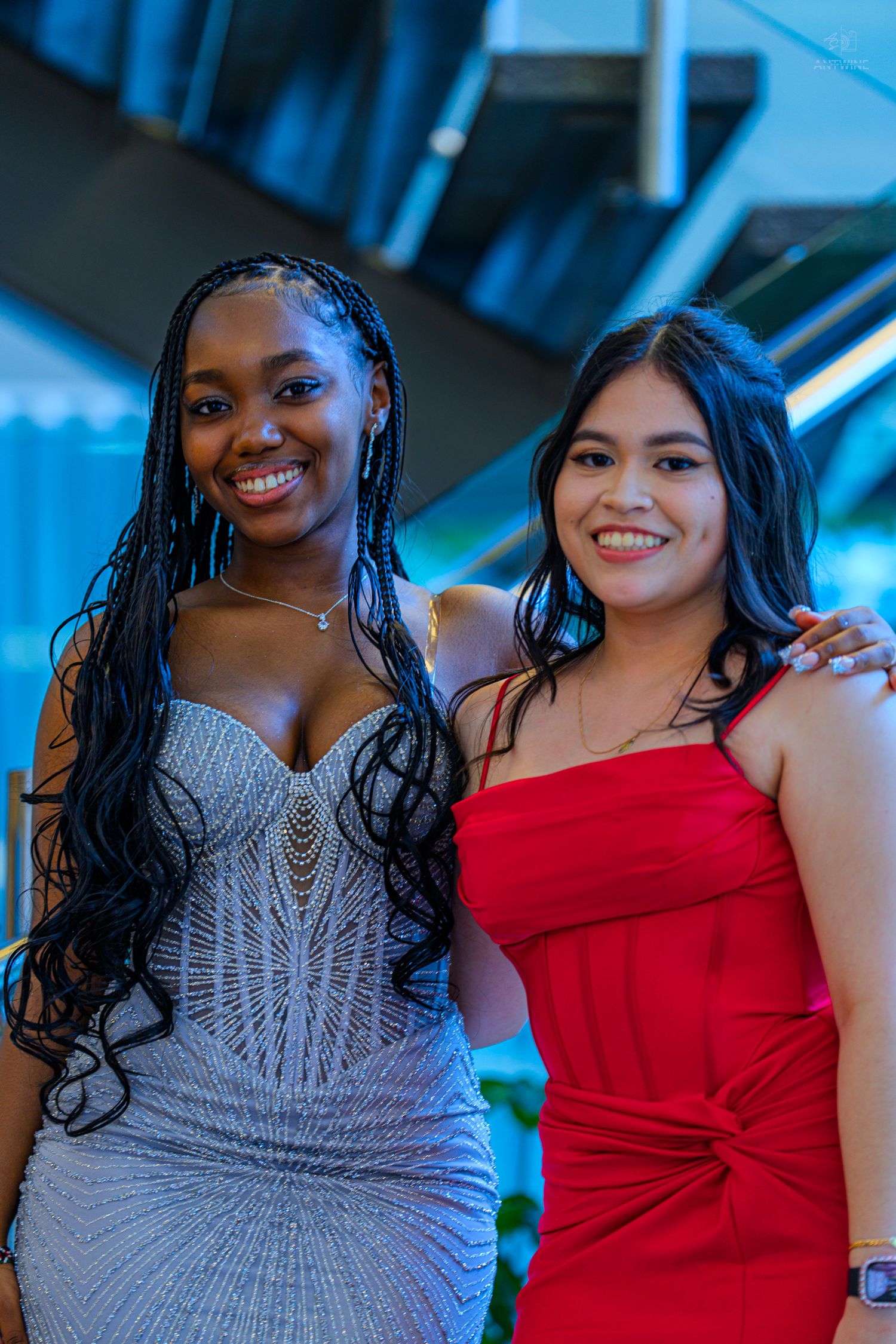 Two smiling young women in elegant formal dresses, one silver and one red, posing together at a prom event.