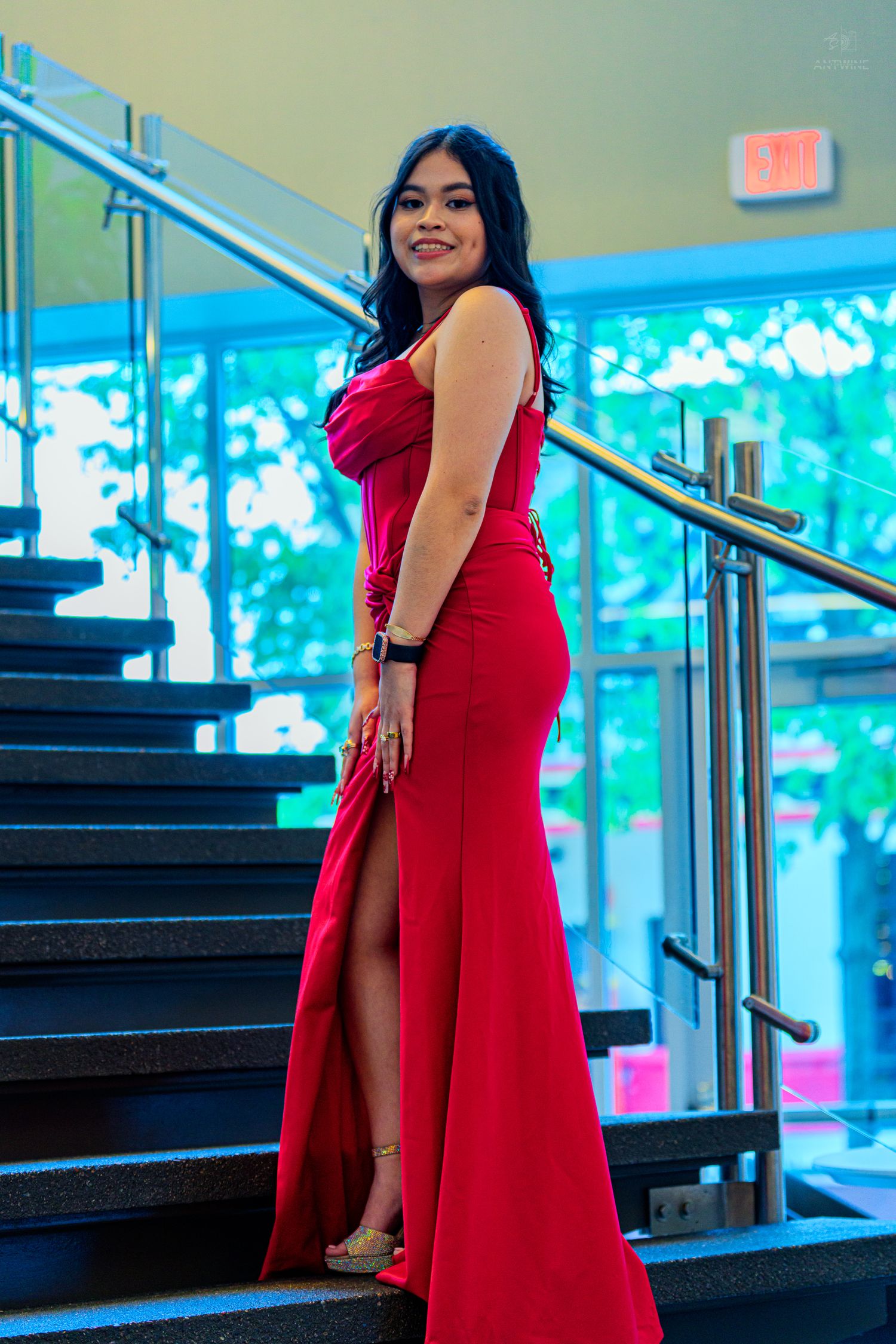 Smiling woman in red formal gown standing on dark staircase with glass railing.