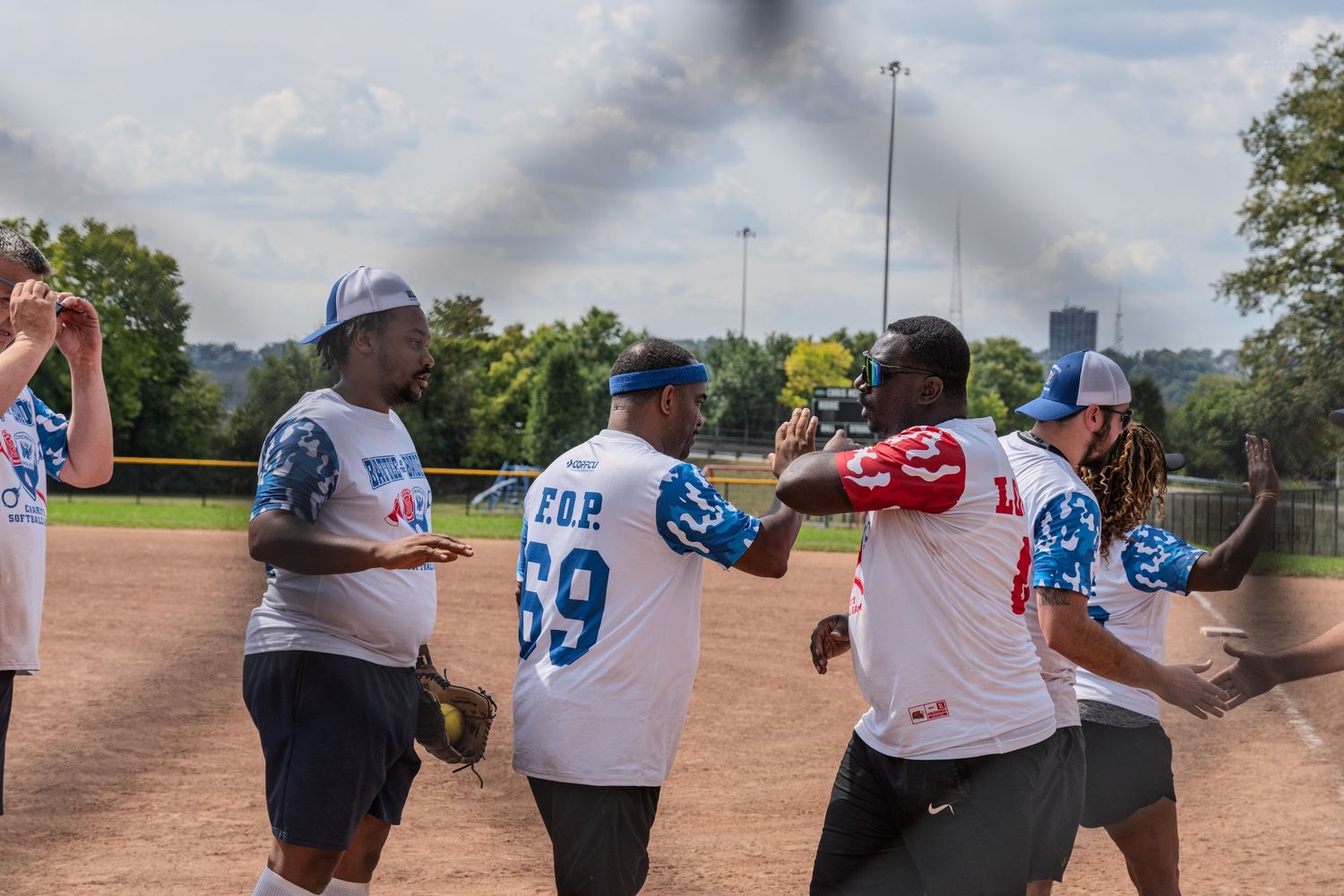 Players in blue and white uniforms celebrate on a softball field during a sunny outdoor game.