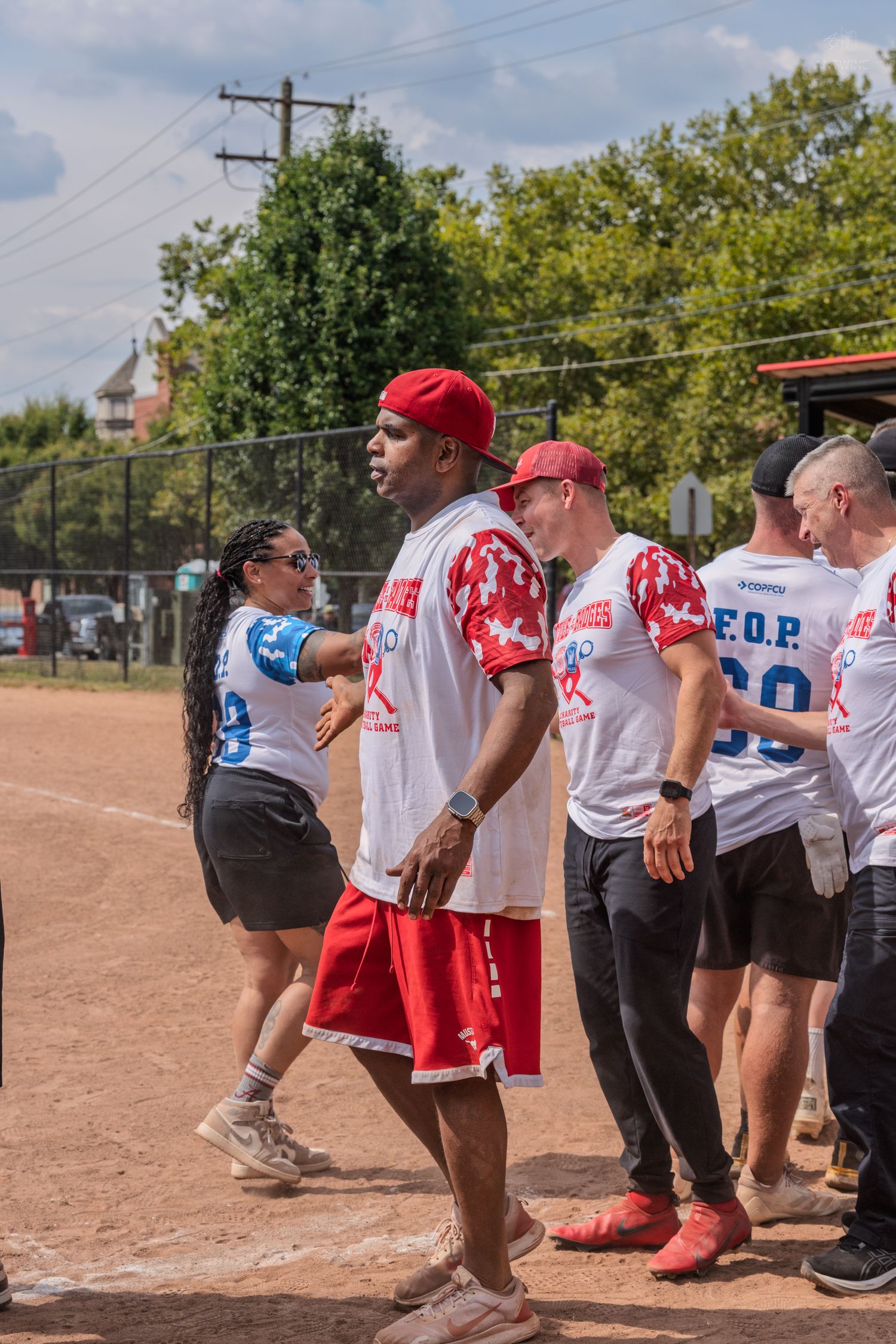 Athletes in red and white softball uniforms socializing on a baseball field during a sunny outdoor charity game.